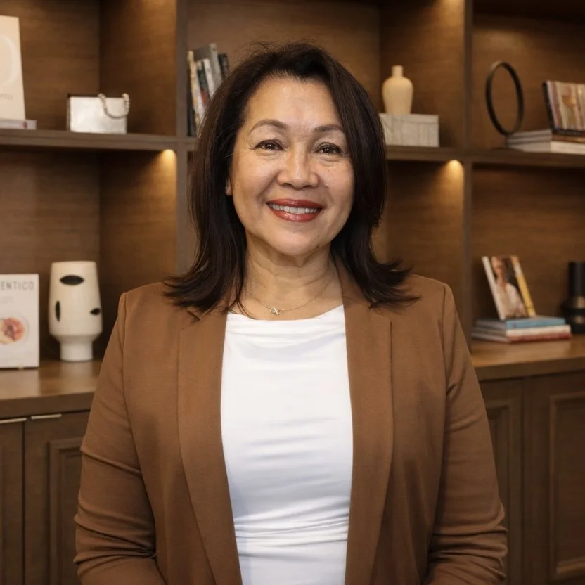 A woman with shoulder-length dark hair and a bright smile, wearing a light brown blazer over a white top, standing in front of wooden shelves filled with books and decorative items.
