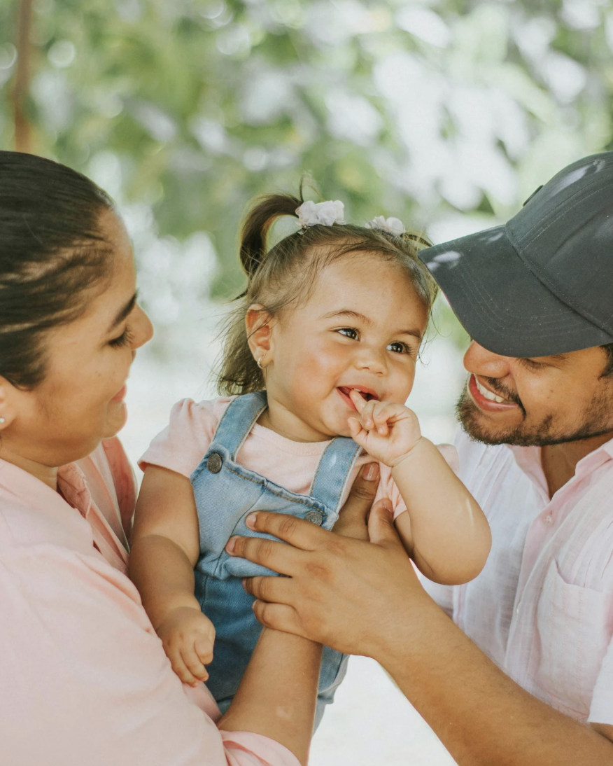 A smiling young girl with a ponytail and wearing overalls is being held by her mother and father; she is happily licking her finger.