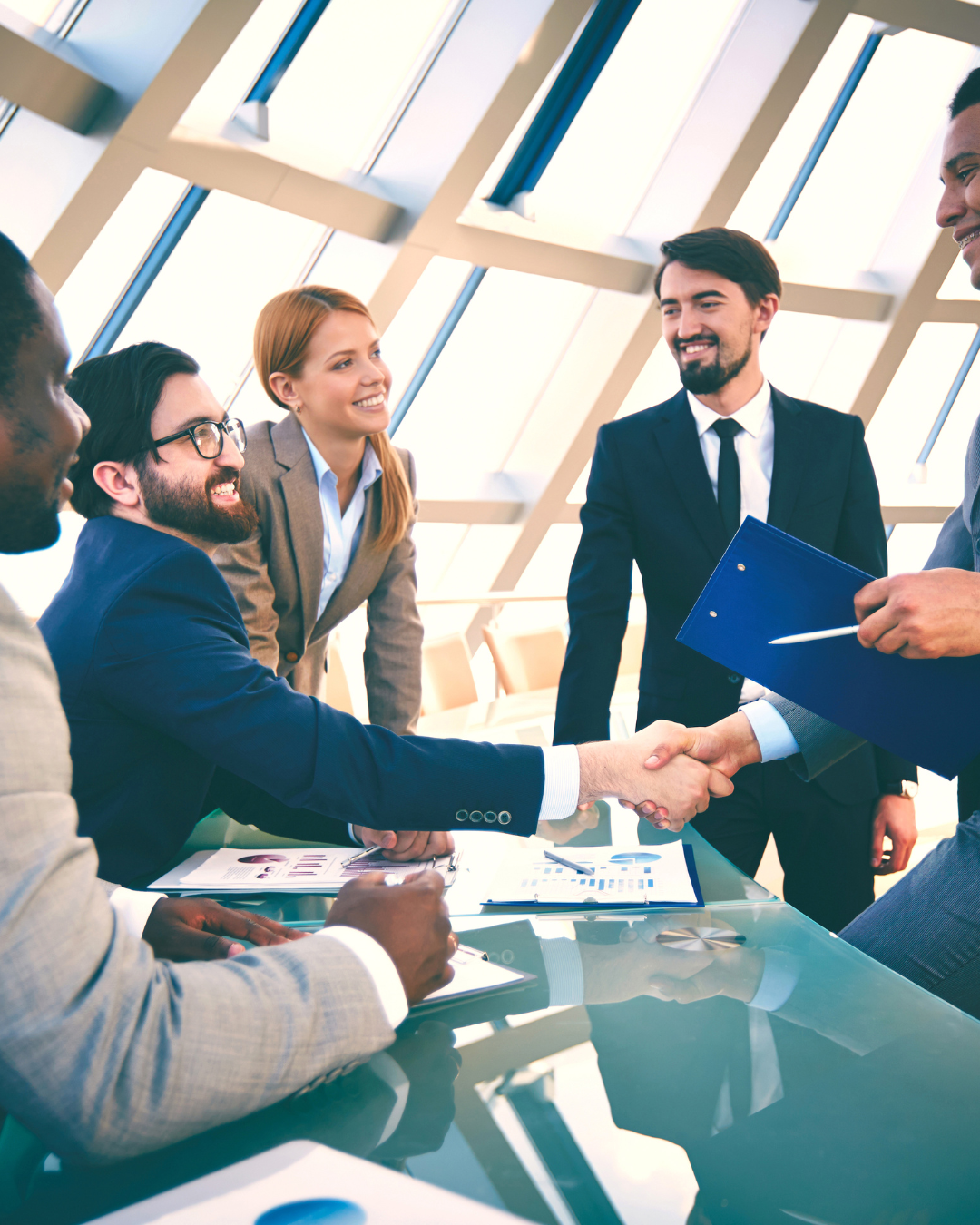 Business professionals in a meeting room shaking hands and smiling, with documents and electronic devices on the table, in a modern office with large windows.