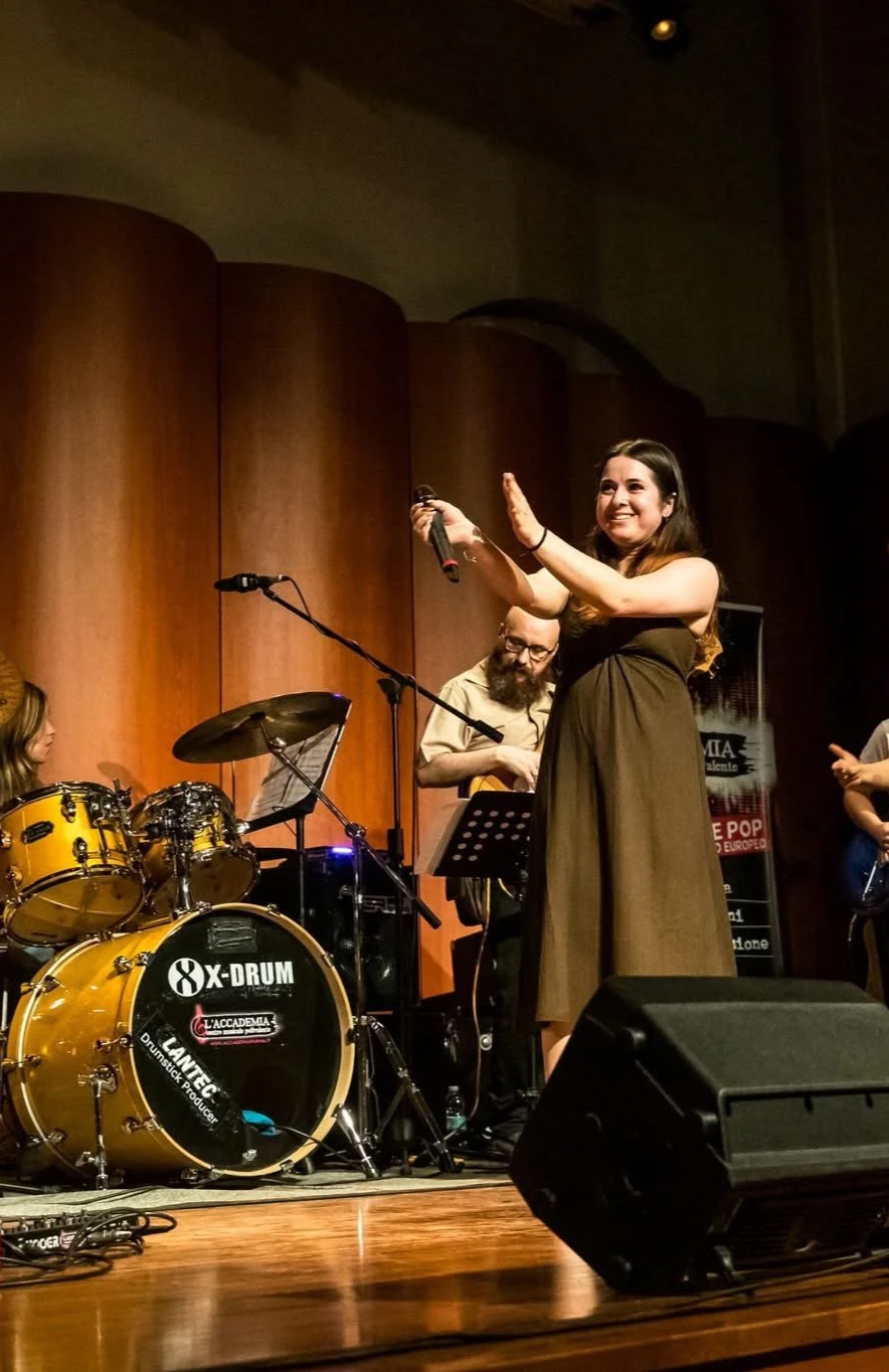 A woman performing on stage with a band, holding a microphone, smiling, and raising her hand. The band includes a drummer with a yellow drum set and a guitarist with a beard, wearing glasses. The stage has a wooden backdrop and musical equipment.