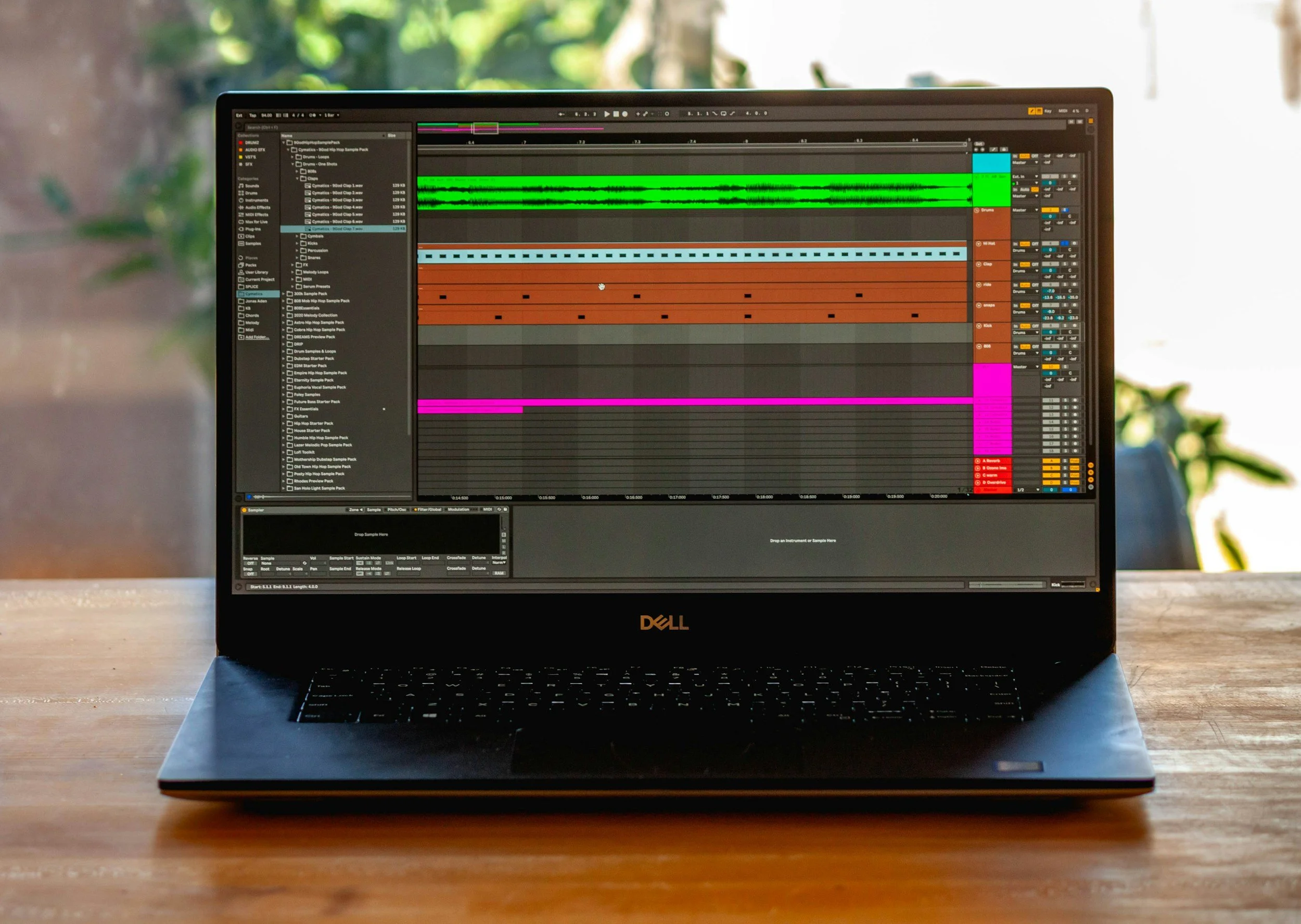 Laptop with digital audio workstation software open, displaying multiple colorful tracks and waveforms, placed on a wooden table with a blurred background of greenery.