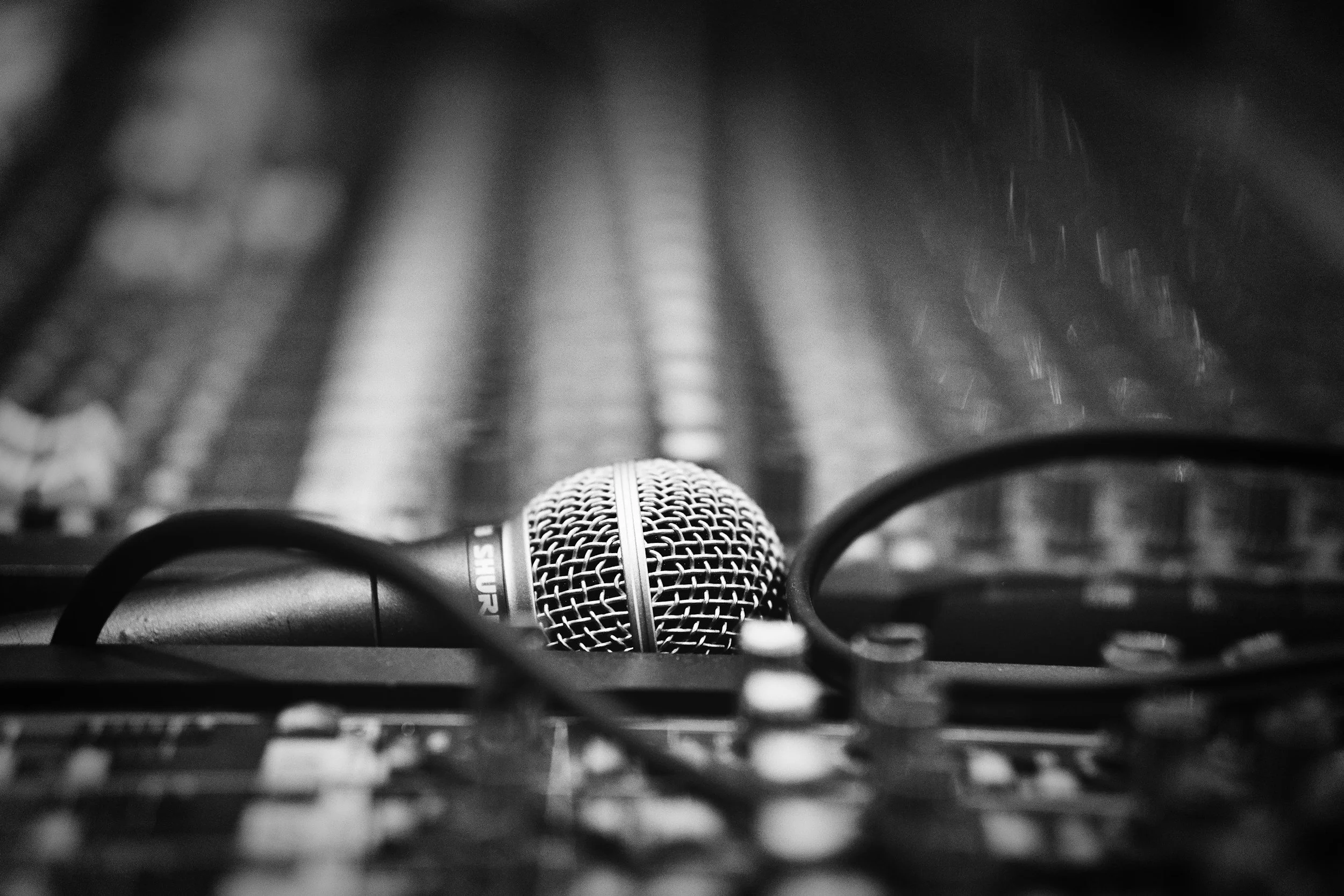 A microphone viewed from below on a music studio desk, with blurred background of audio equipment and a dark atmosphere.