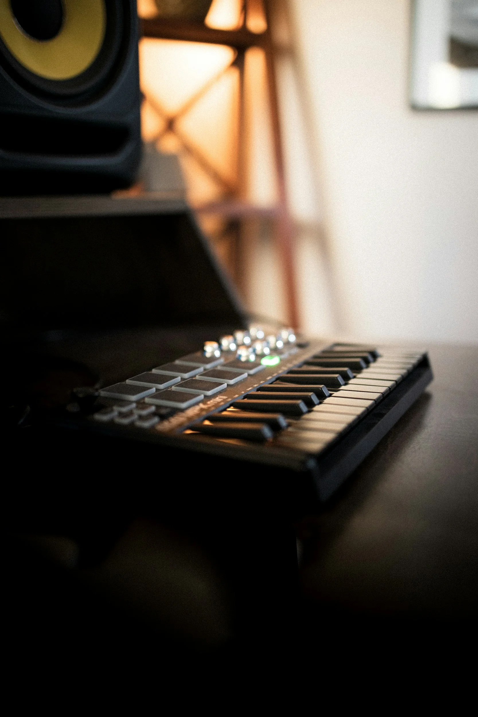 Close-up of a modern digital keyboard with black and white keys, and a speaker on the left side, with blurred warm lighting and furniture in the background.