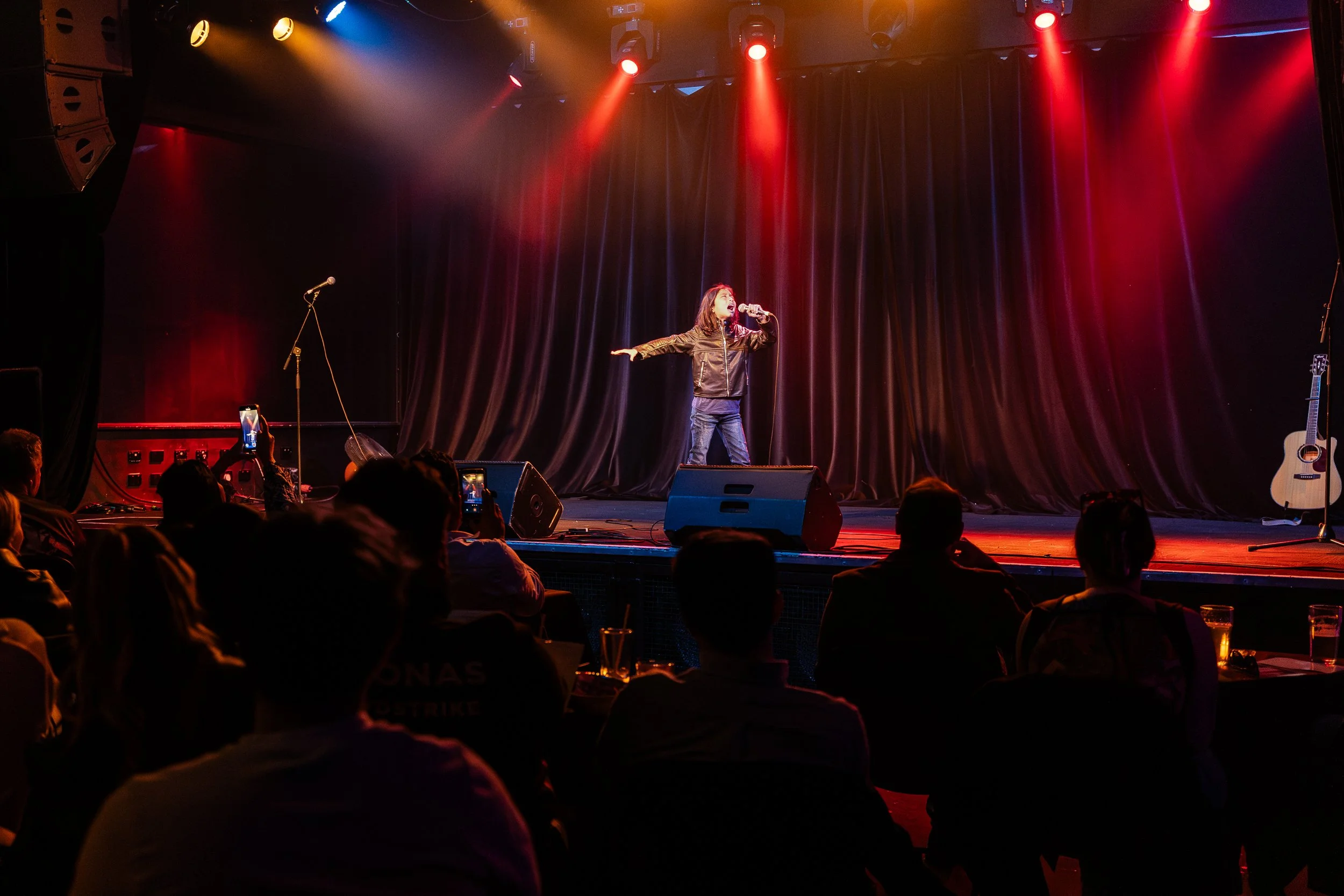 A performer singing on stage with colorful stage lights shining down, accompanied by a guitar on the side, and an audience watching in front.