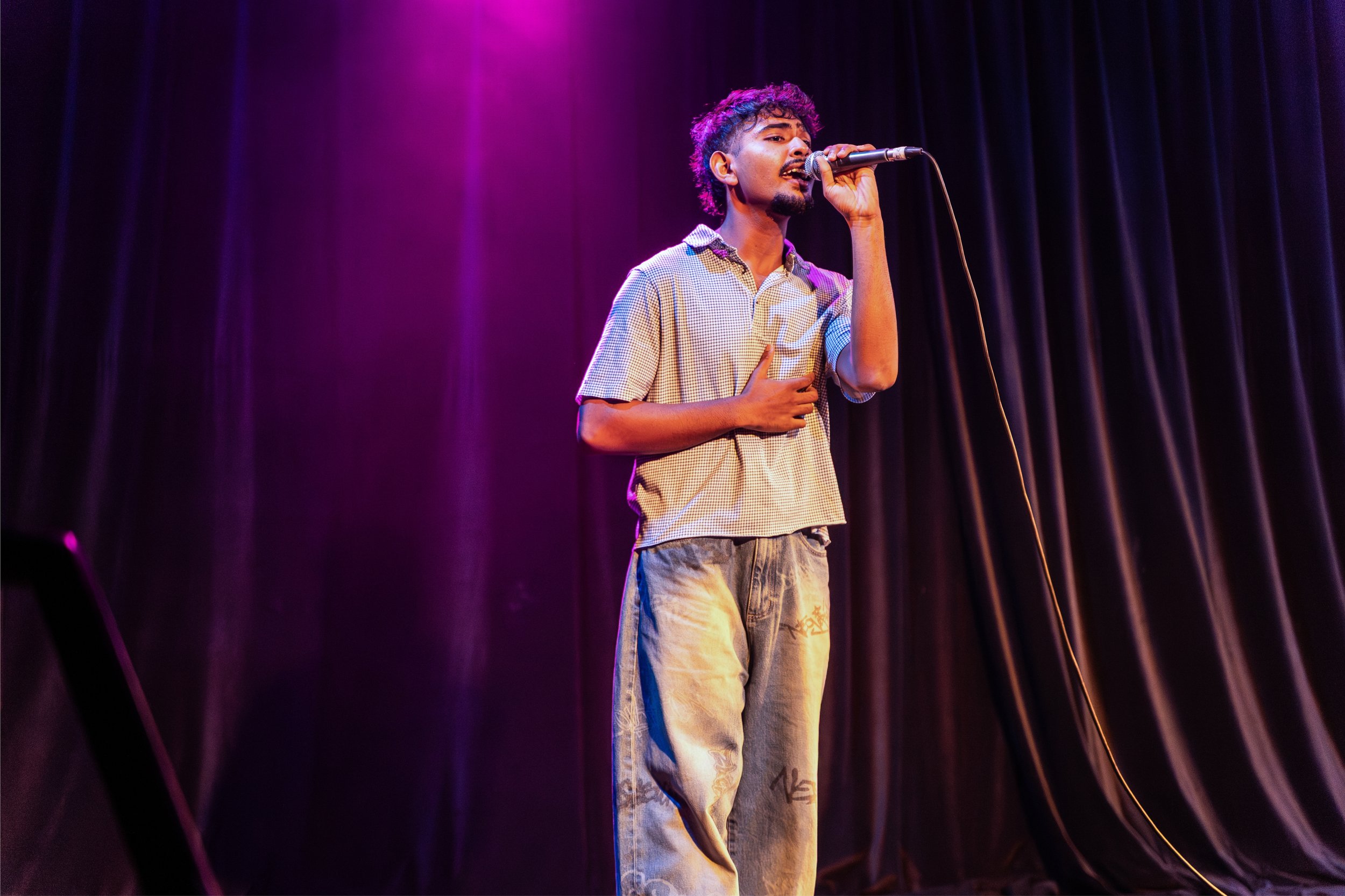 A young man with curly hair and a mustache singing into a microphone on stage, wearing a checkered shirt and patterned jeans, with purple stage lighting and dark curtains in the background.