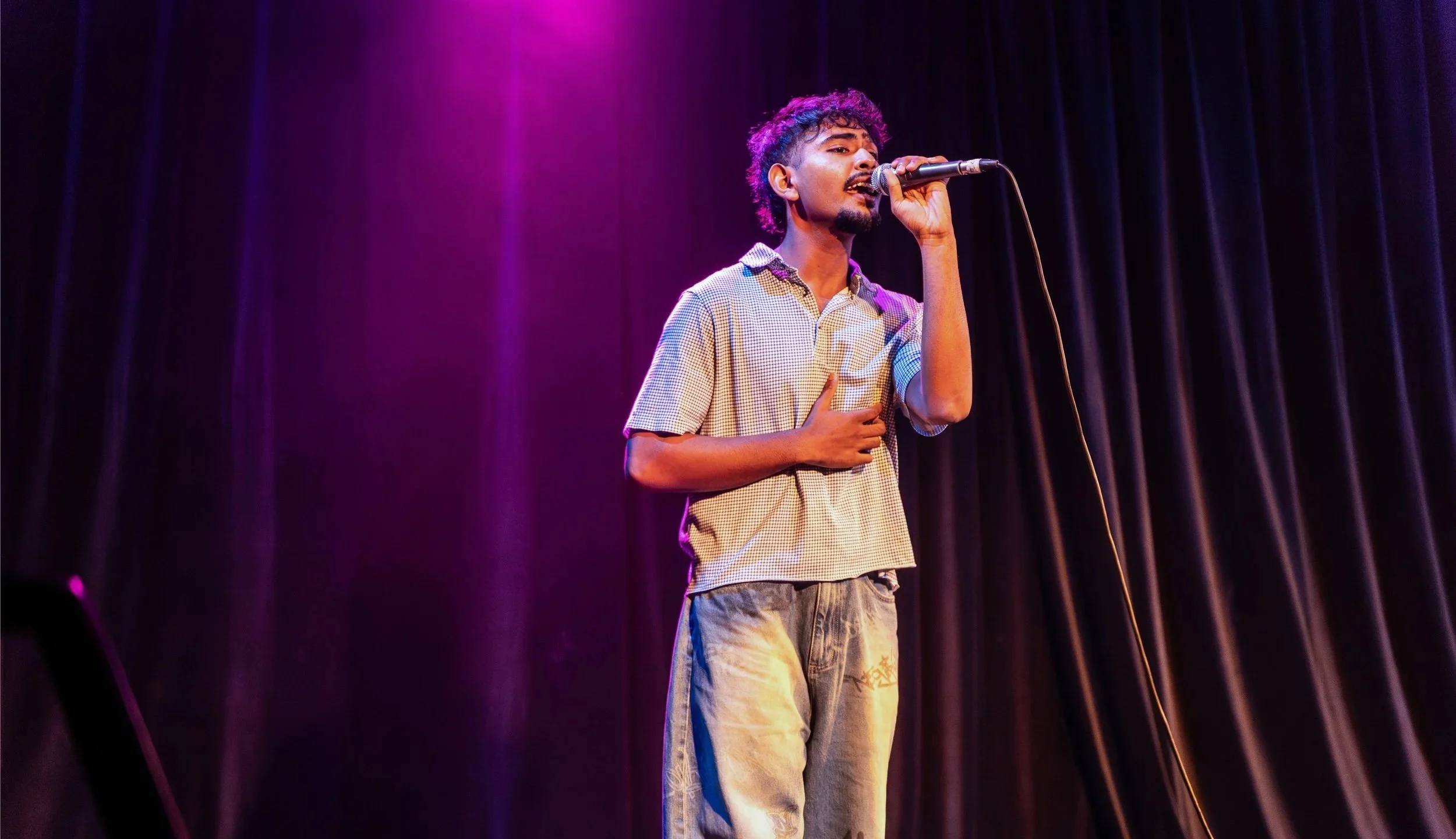 Young man with curly hair singing into a microphone on stage with purple and pink lighting and dark curtains in the background.