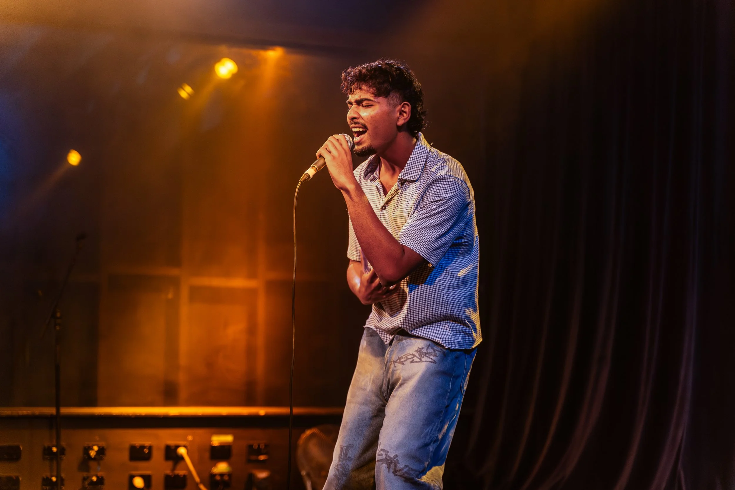 Young man singing passionately into a microphone on stage, dressed in a checkered shirt and jeans, with stage lights illuminating him in yellow and orange hues.