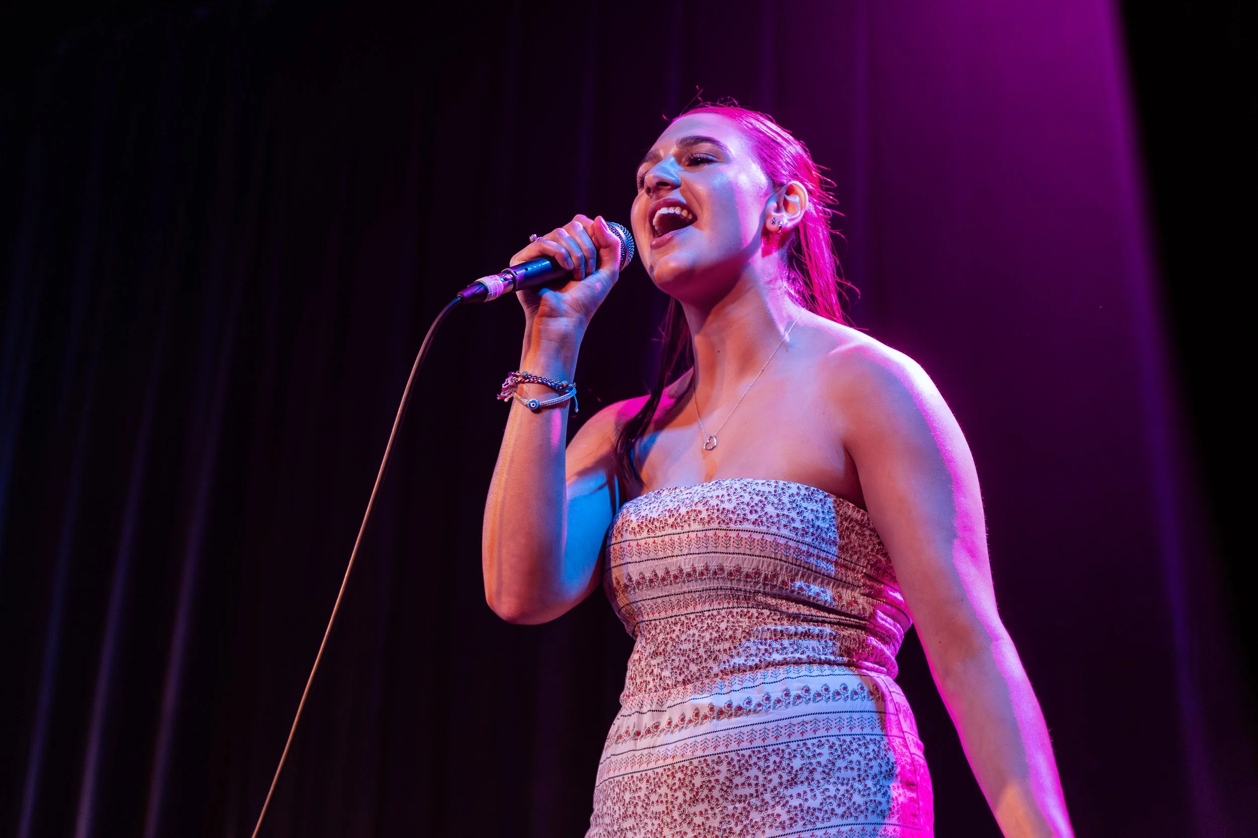 A woman singing into a microphone on stage with purple lighting.