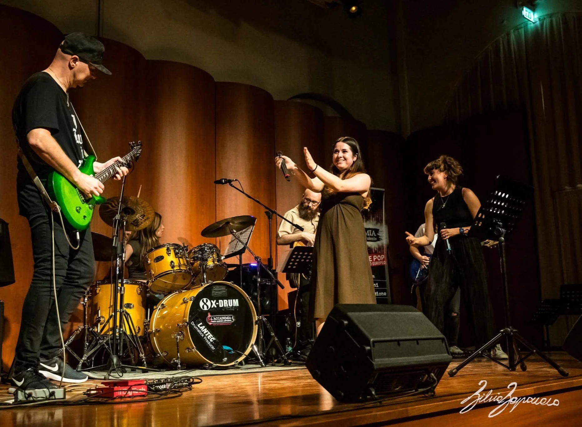A group of musicians performing on stage, including a woman singing into a microphone, a guitarist playing, a drummer, and a woman singing or speaking with a microphone in her hand. The stage has wooden paneling and various musical instruments.