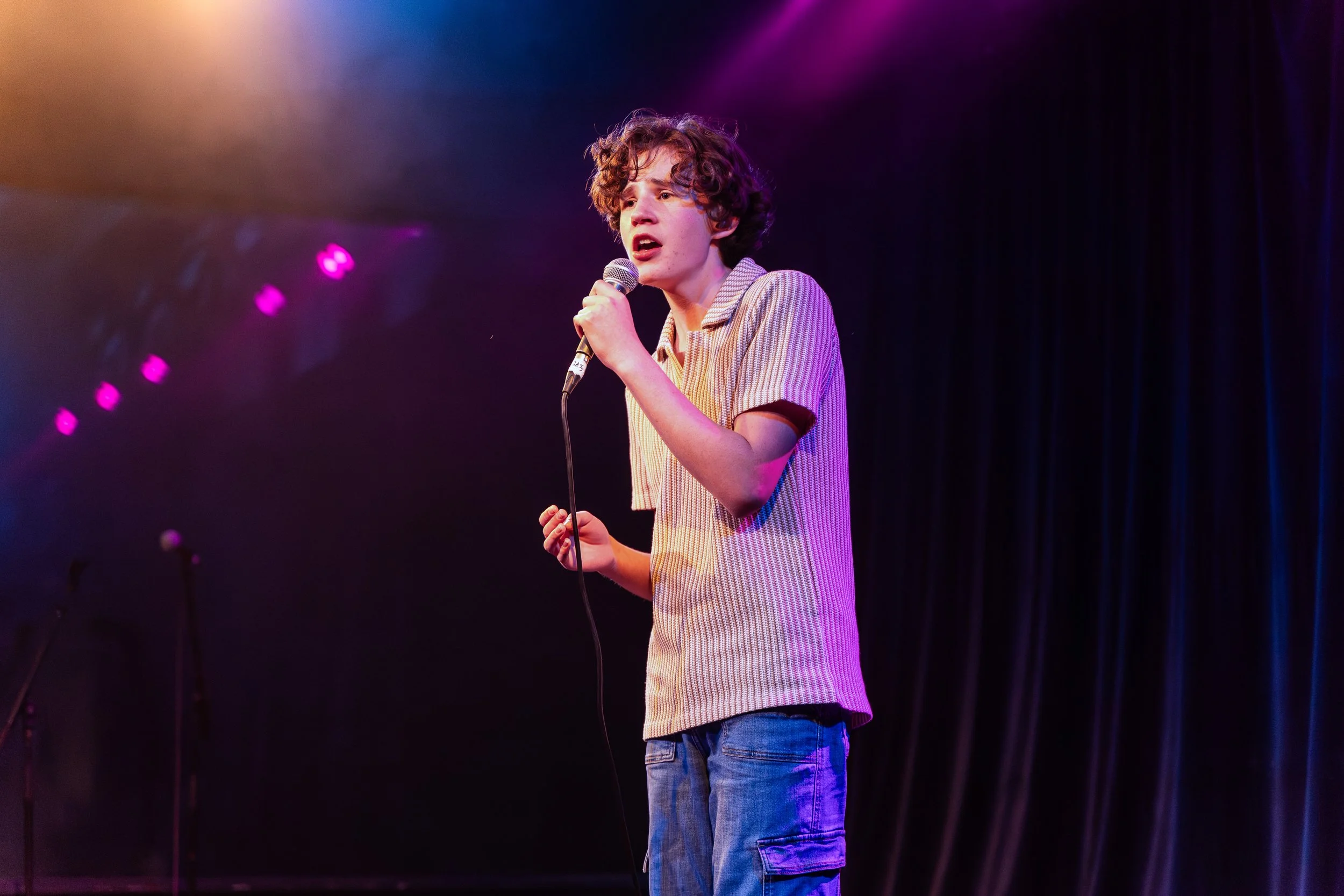 A young man with curly brown hair singing into a microphone on stage, illuminated by purple and pink stage lights with dark curtains in the background.