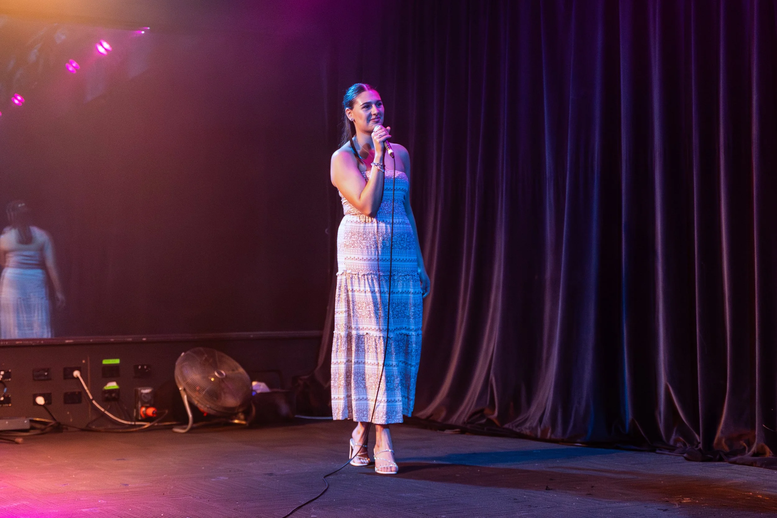 A woman standing on stage, holding a microphone, dressed in a long patterned dress and high heels, with dark hair pulled back, under colorful stage lighting, with purple curtains in the background.