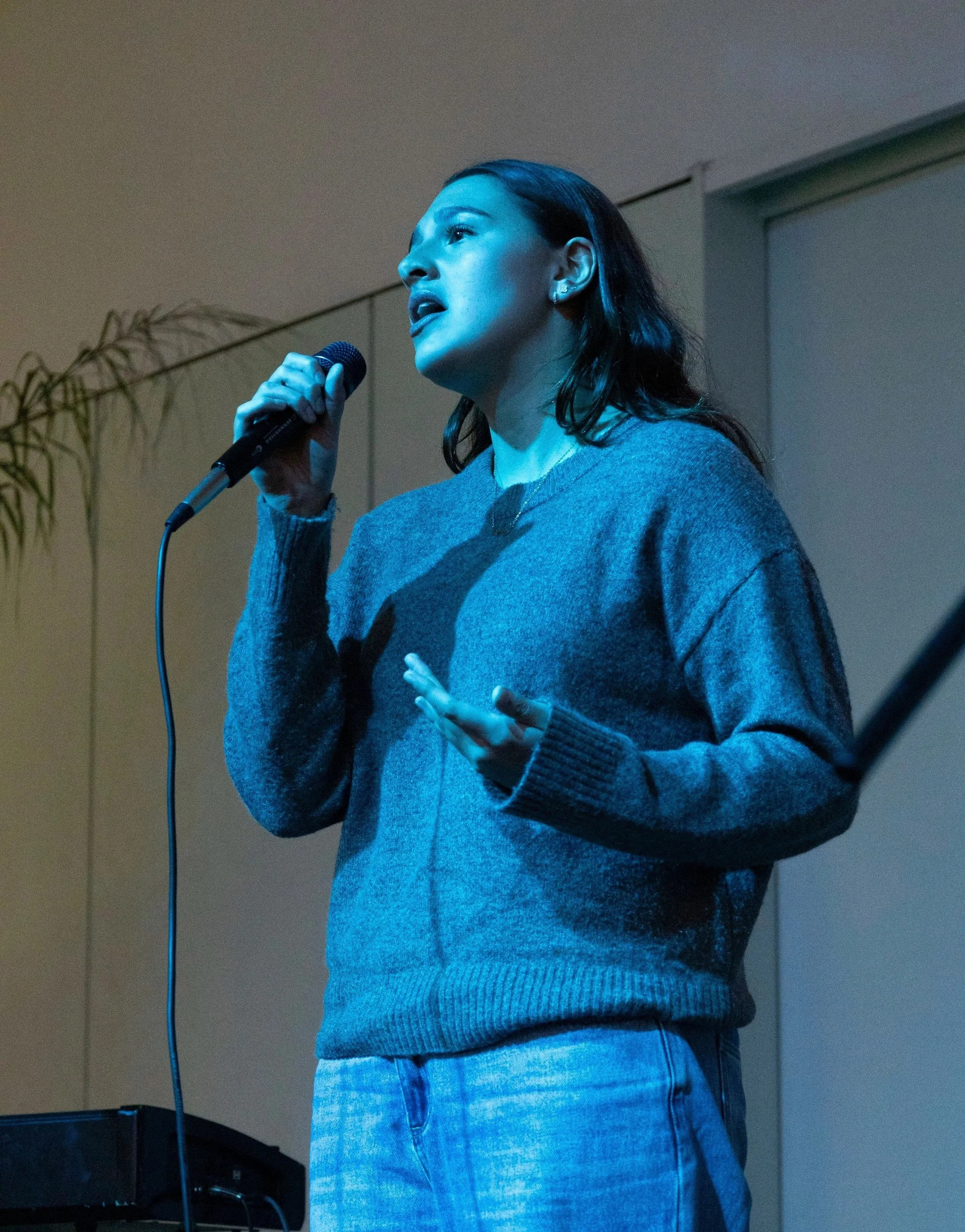 A woman with dark hair singing into a microphone at an indoor event, wearing a gray sweater and a necklace, with a plant and a window in the background.