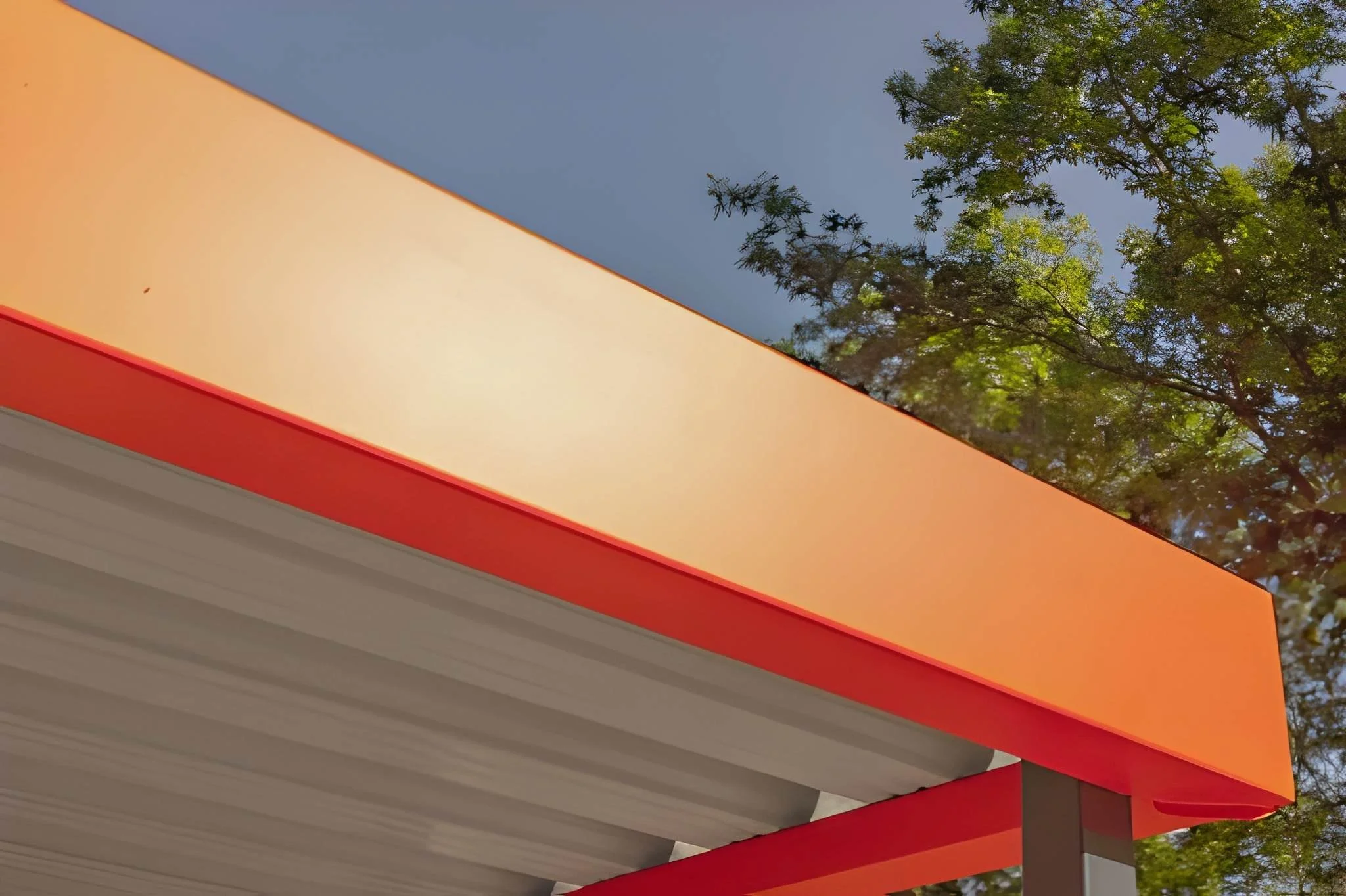 Close-up of a building corner with orange and red exterior panels, with a background of blue sky and green tree branches.