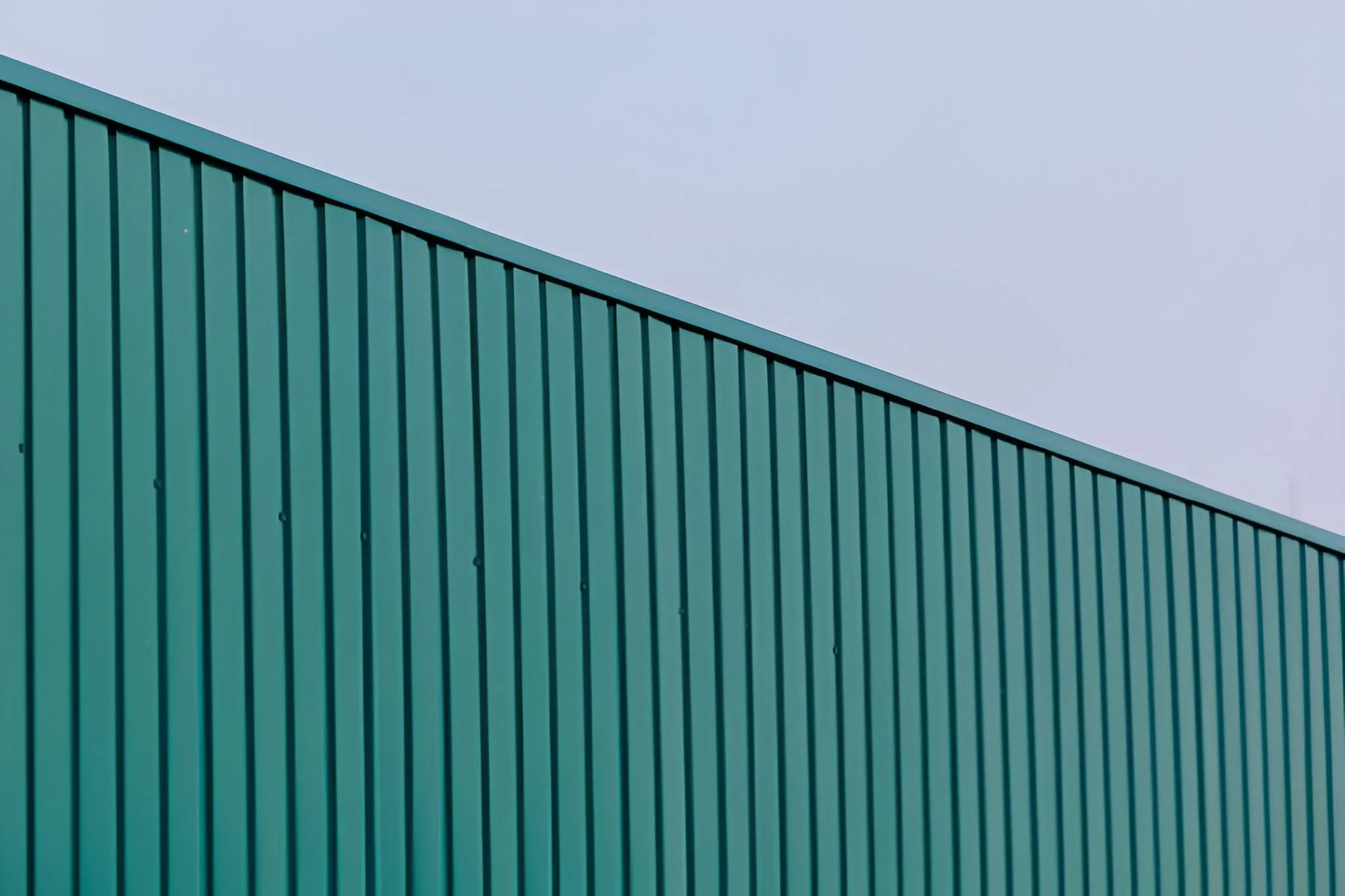 Close-up of a teal metal building with vertical ridges, corner visible, under a cloudy sky.