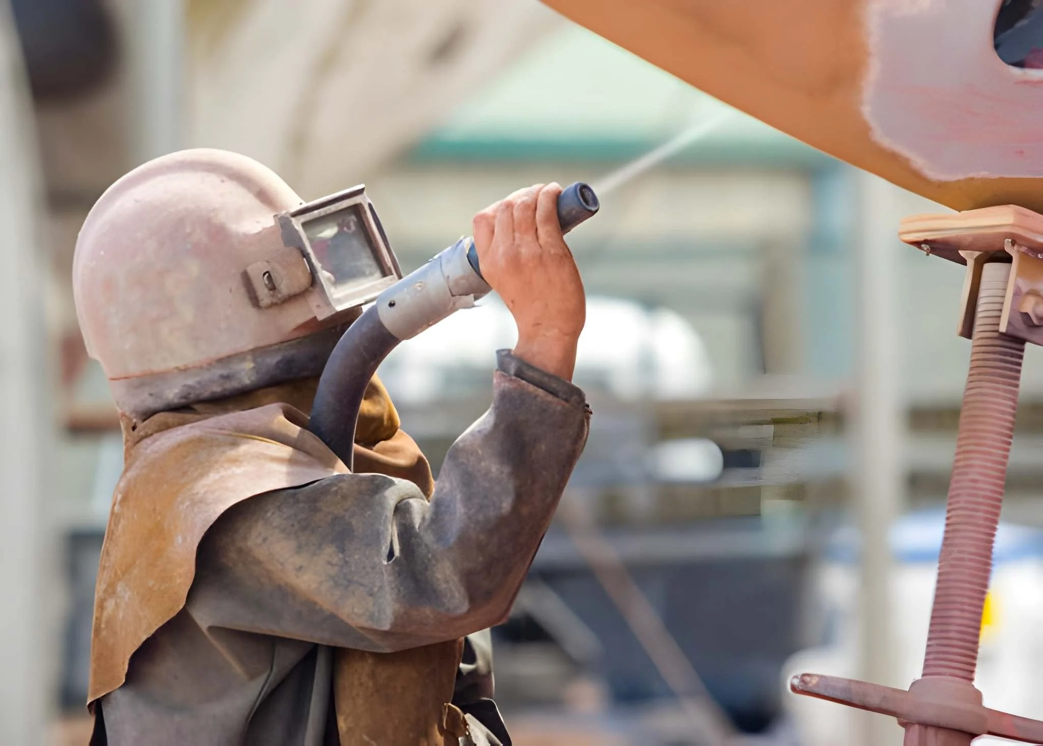 A worker with a dust-covered helmet and protective jacket welding or grinding in an industrial workshop.