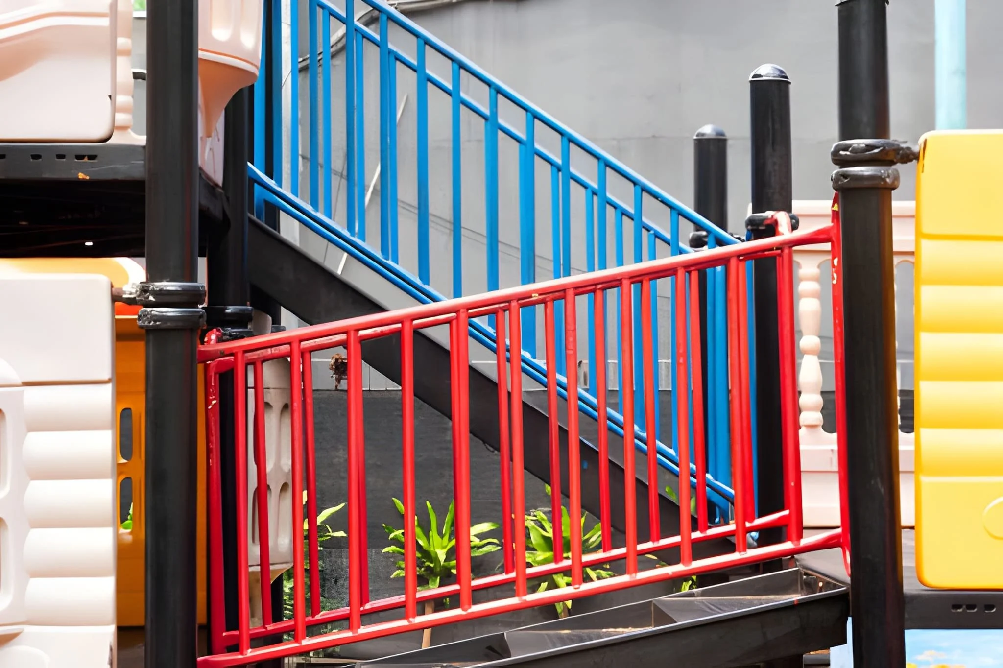 Colorful playground stairs with red, blue, yellow, and black railings and steps, and some green plants at the bottom.