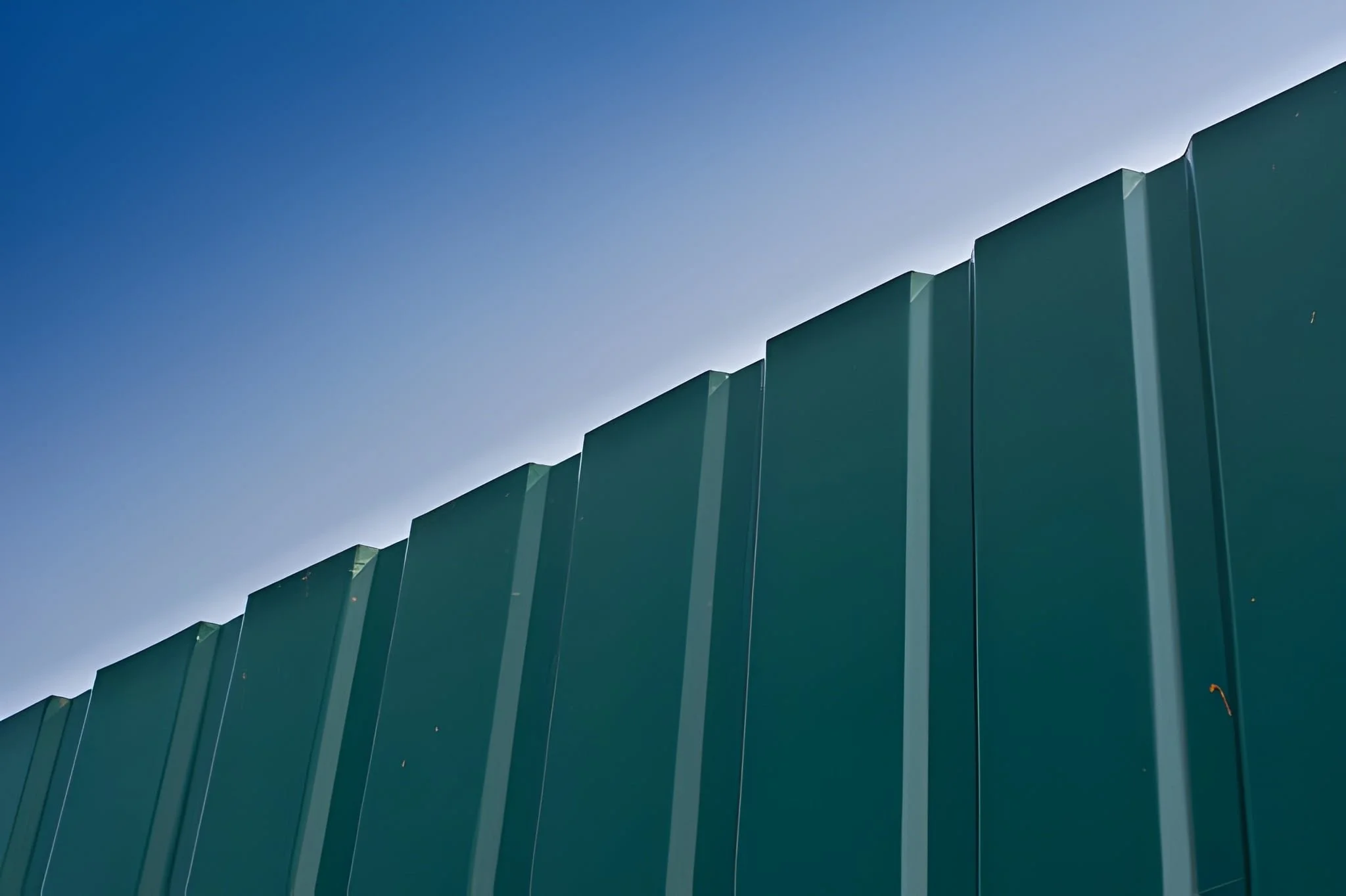 Close-up of a green metal fence against a blue sky with minimal clouds.