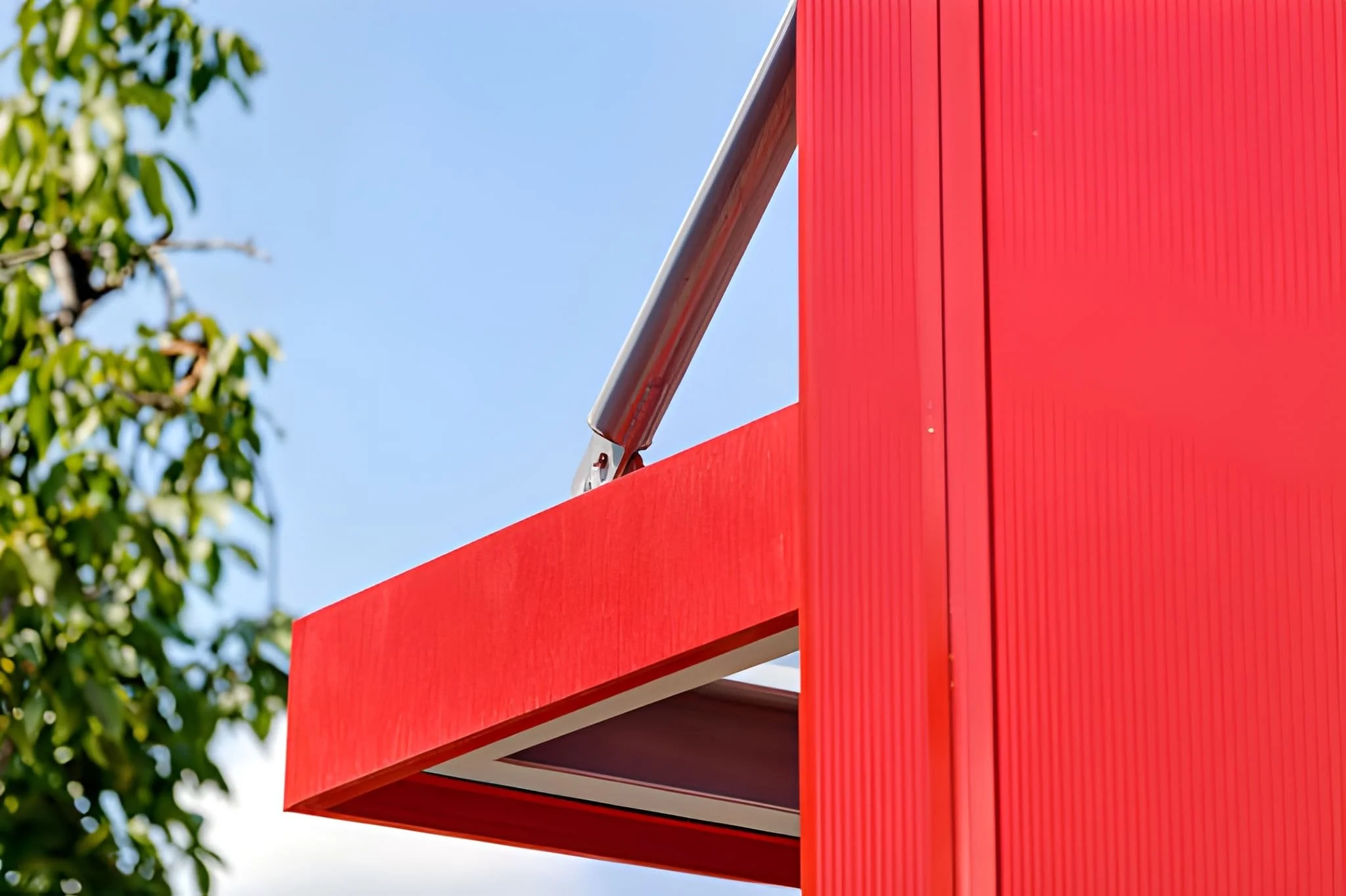 Close-up of a modern red building with a metal railing, a clear blue sky, and green tree leaves on the left.