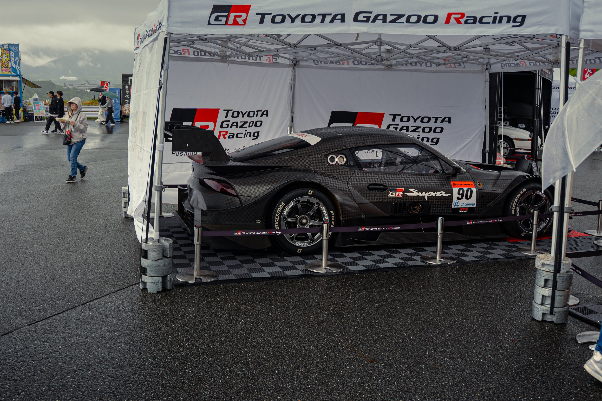 A black race car with a carbon fiber finish on display in a tent with Toyota Gazoo Racing branding at a racing event. The car has a large rear wing, racing tires, and is marked with the number 90.