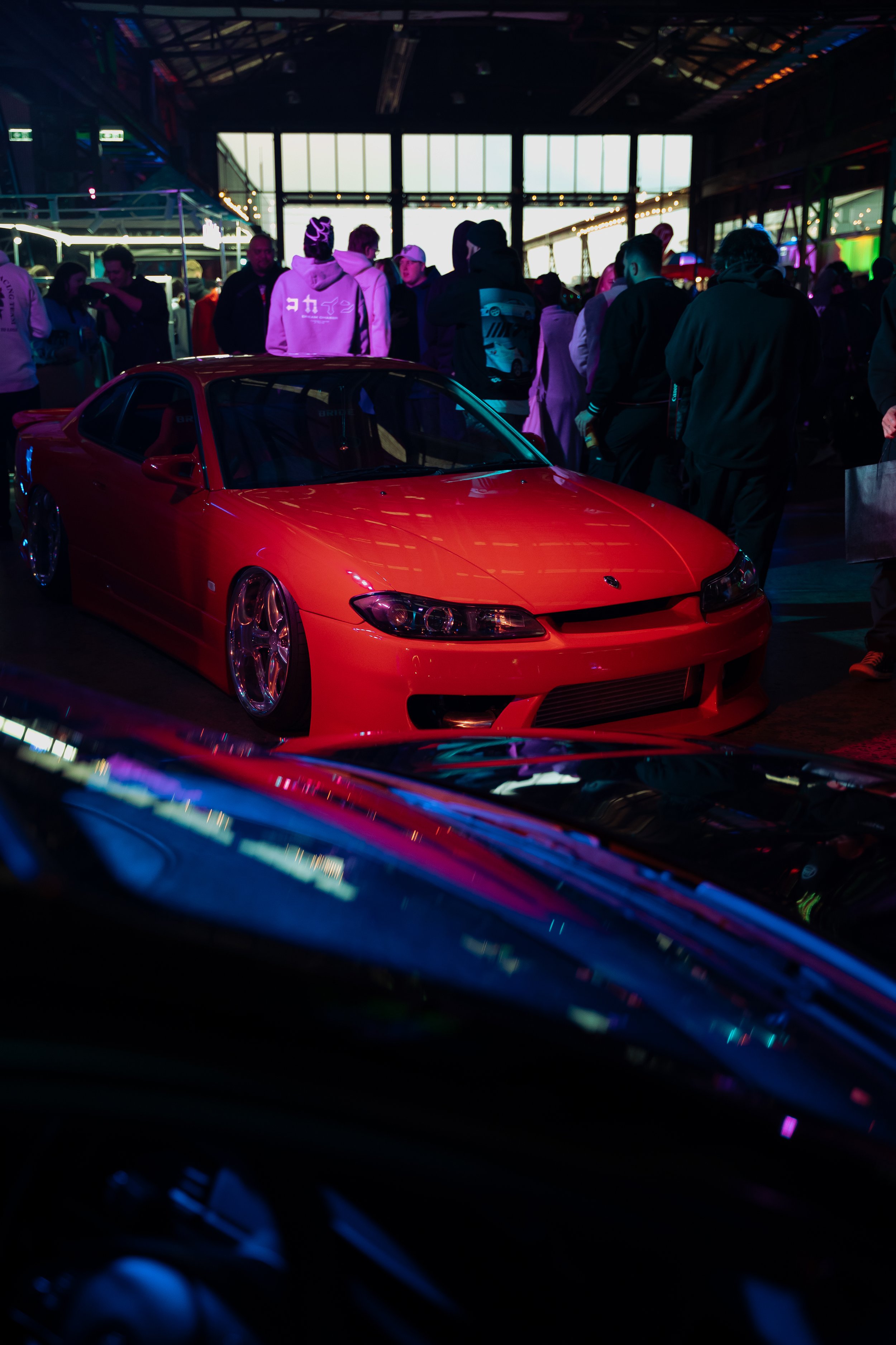 Red sports car on display at an indoor car show with people gathered around and colorful lighting.
