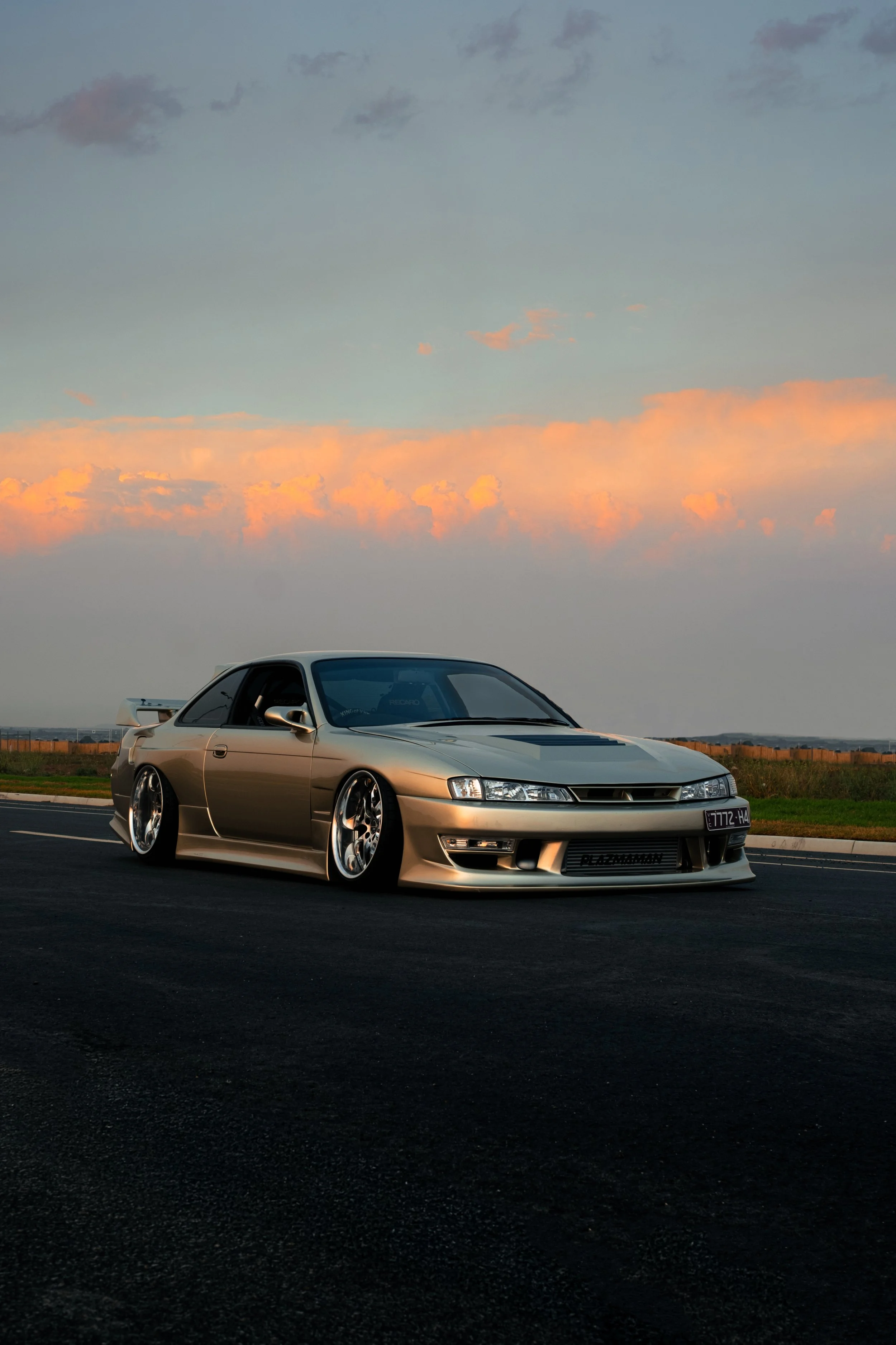 A silver sports car with a wide body kit and custom wheels parked on an empty road at sunset with colorful clouds in the sky.