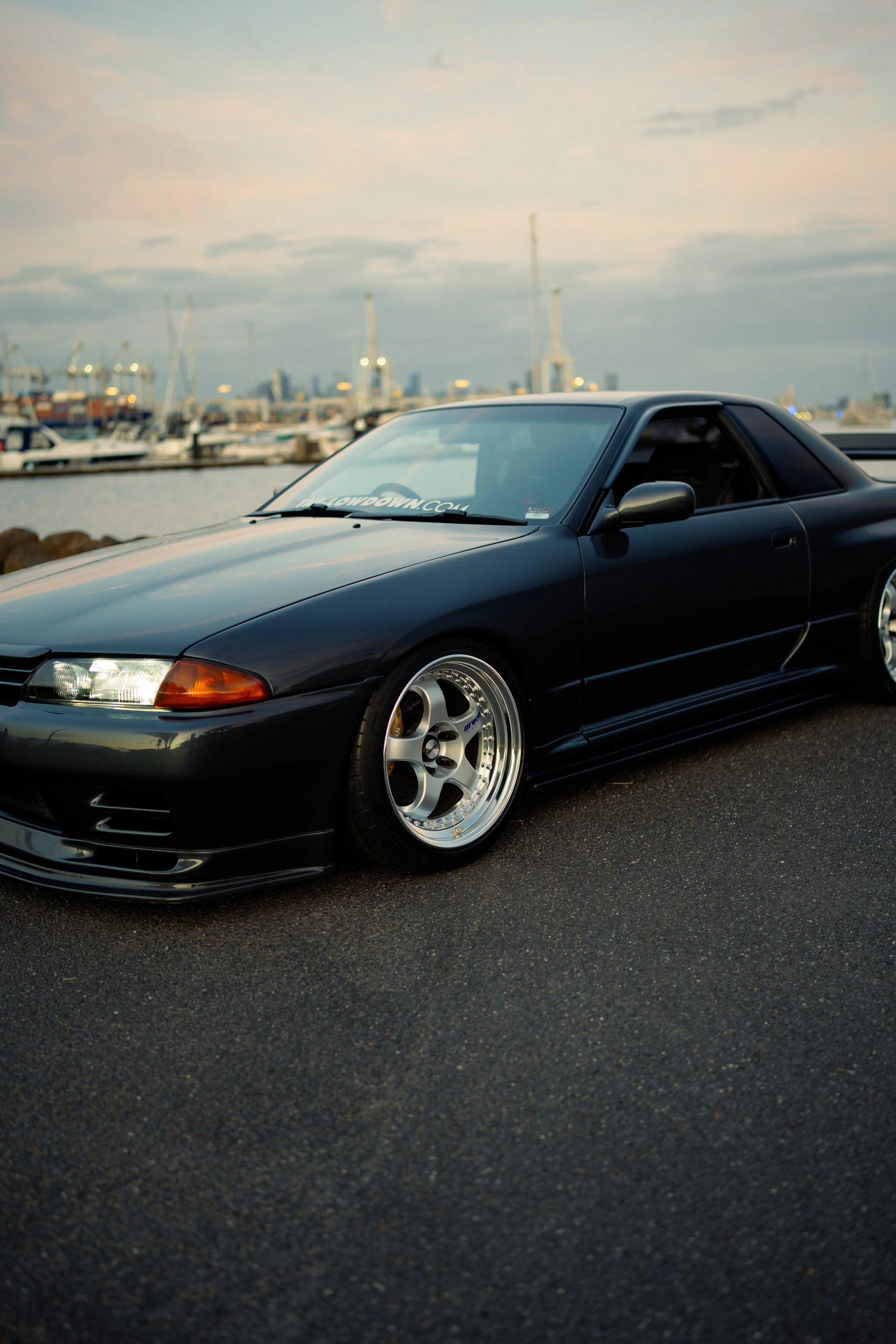 A black sports car with aftermarket wheels parked near a marina with boats and a city skyline in the background during sunset.