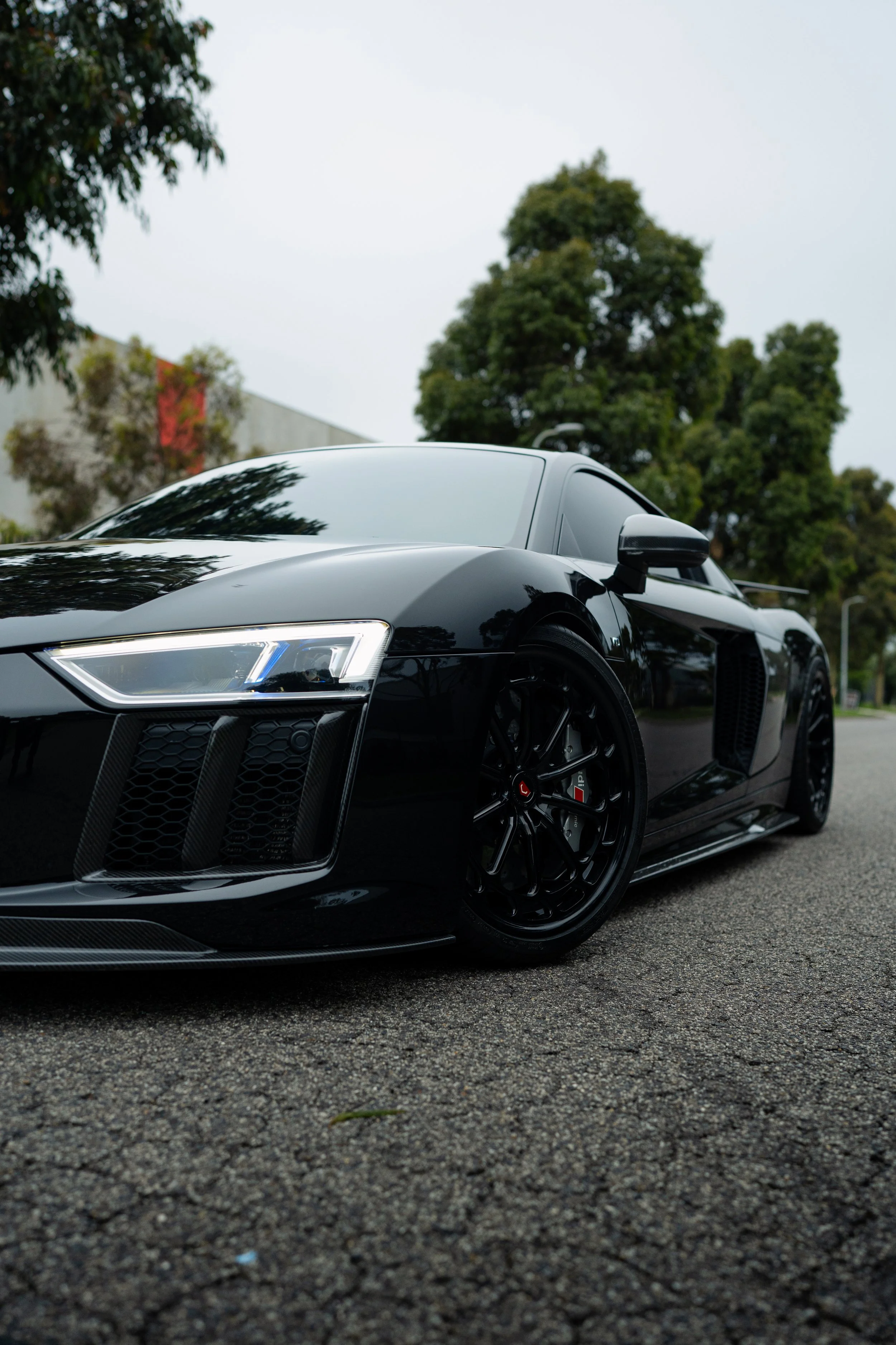 A sleek black sports car parked on an urban street with trees and a building in the background.