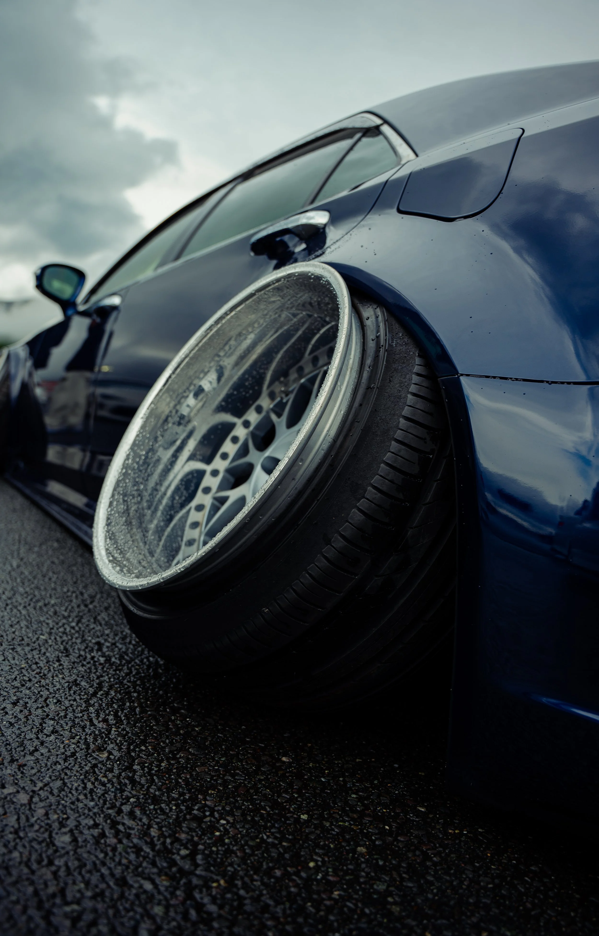 Close-up of a blue sports car with a modified wheel and tire on a wet road, under a cloudy sky.