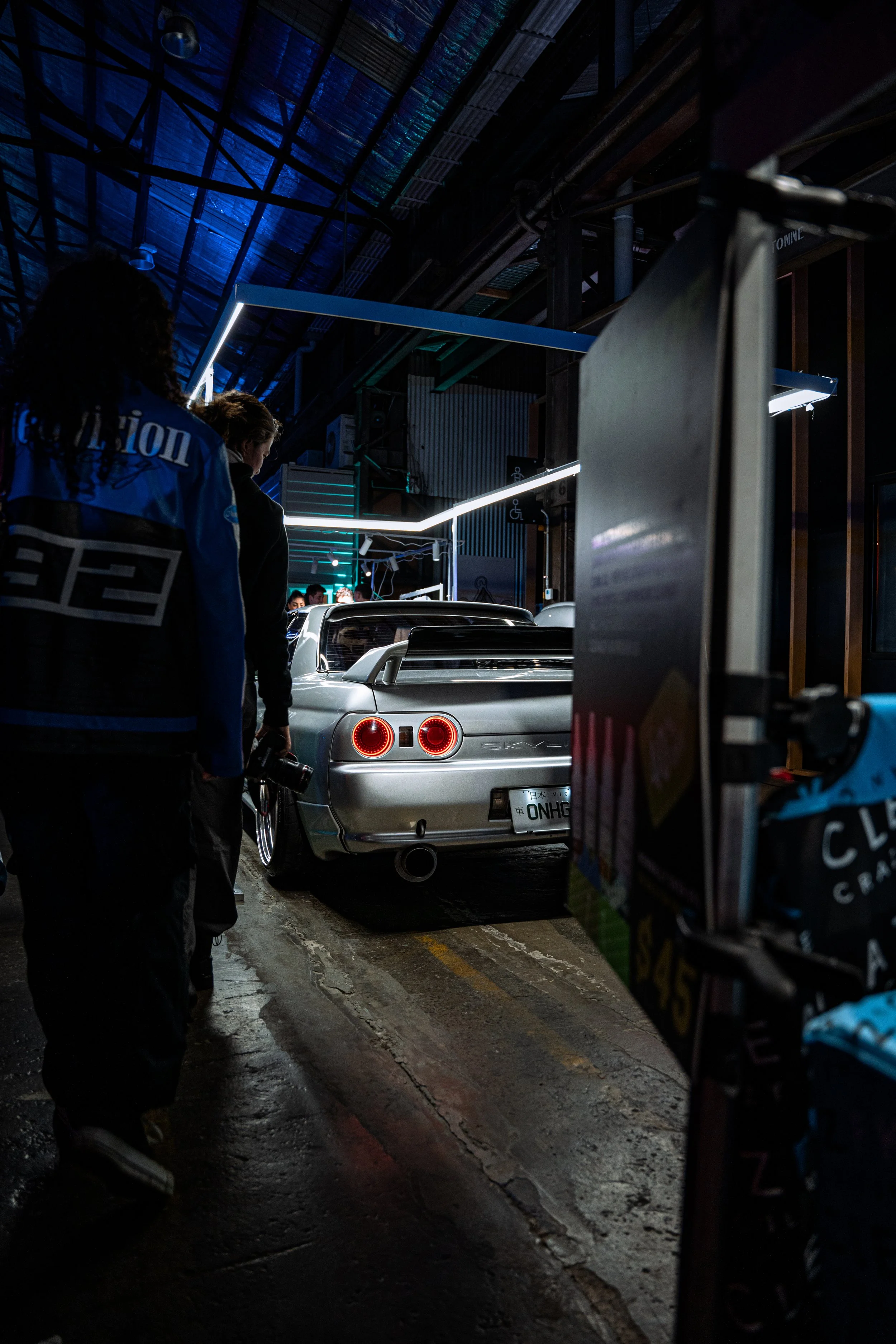 A silver sports car with a rear spoiler parked inside a dimly lit indoor venue, with people walking nearby and neon lighting overhead.