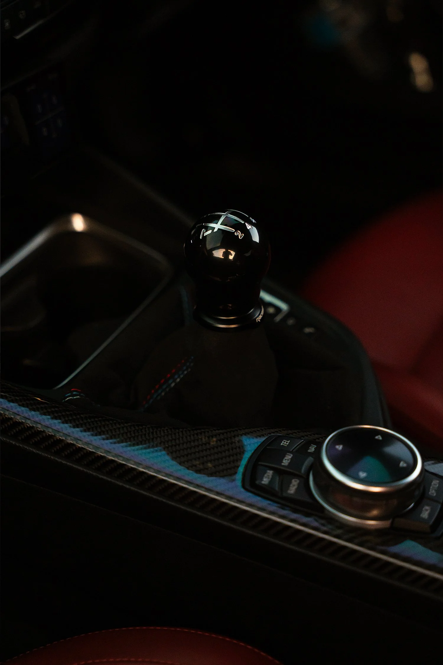 Close-up of a manual gear shift lever in a car interior, with a control dial and buttons nearby, featuring a carbon fiber trim.