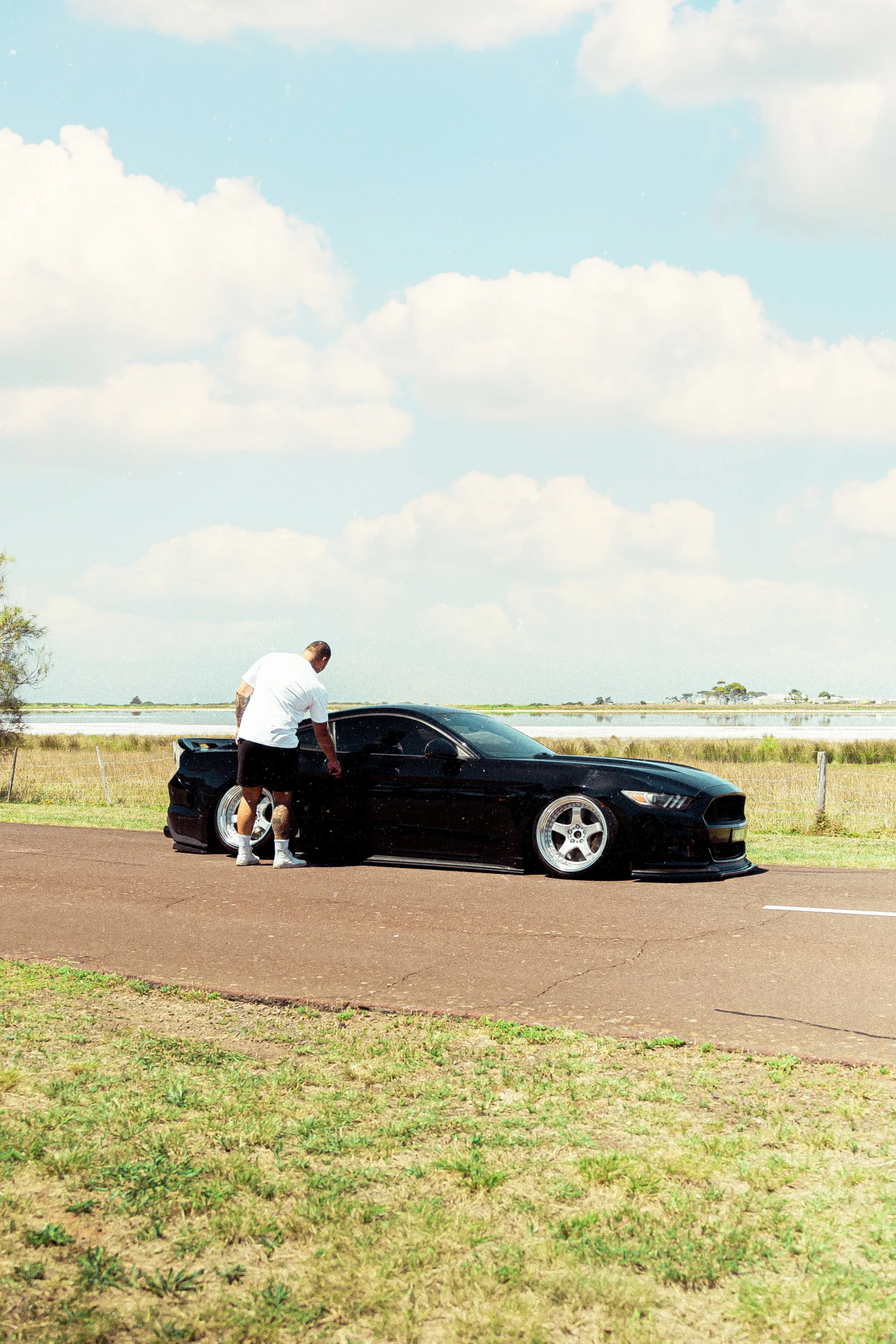 A man in a white shirt and black shorts stands next to a black sports car on an open road with grass and a pond in the background under a partly cloudy sky.