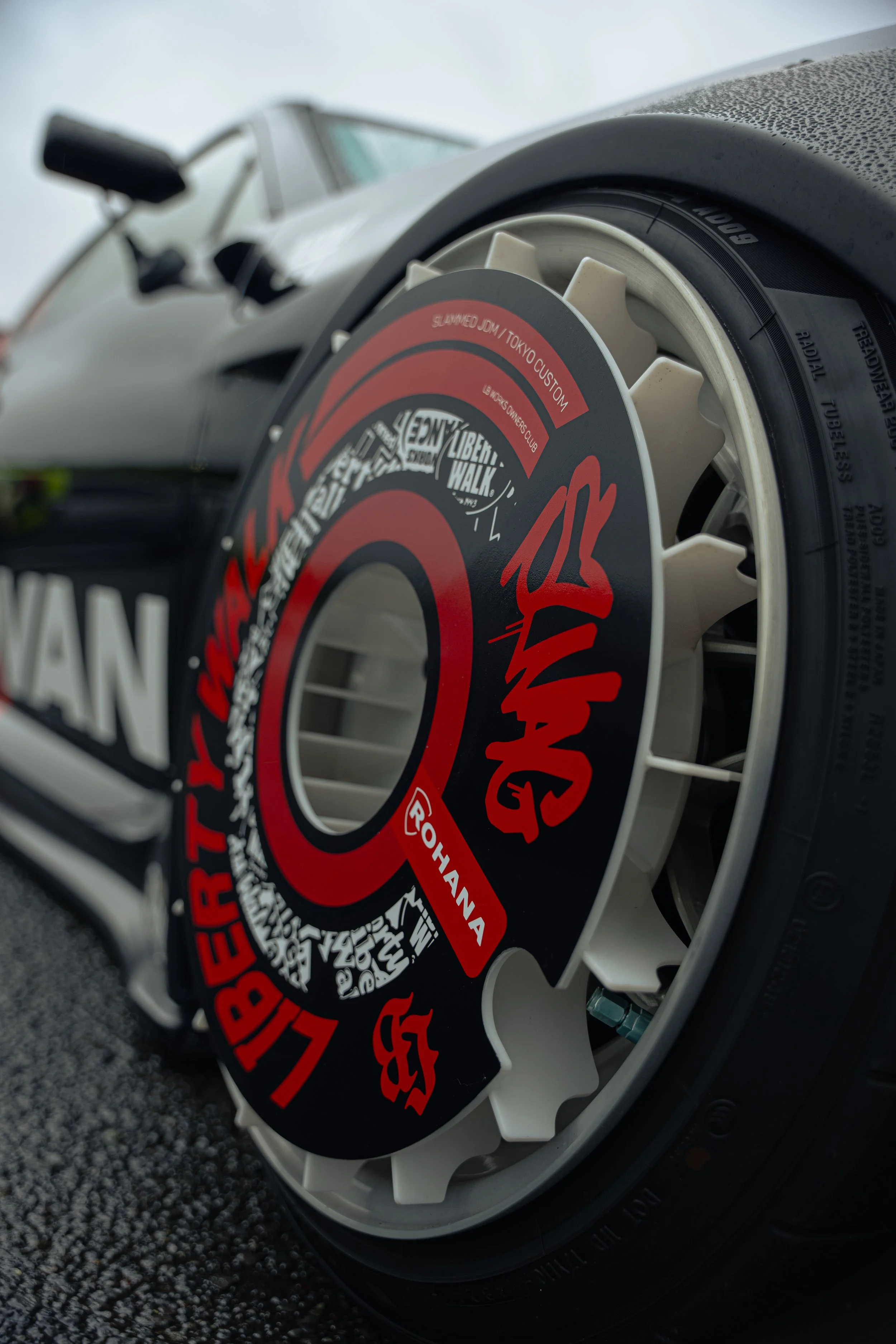 Close-up of a car's wheel with a multi-color wheel cover featuring red, black, and white graphics and text, including a prominent logo or name.