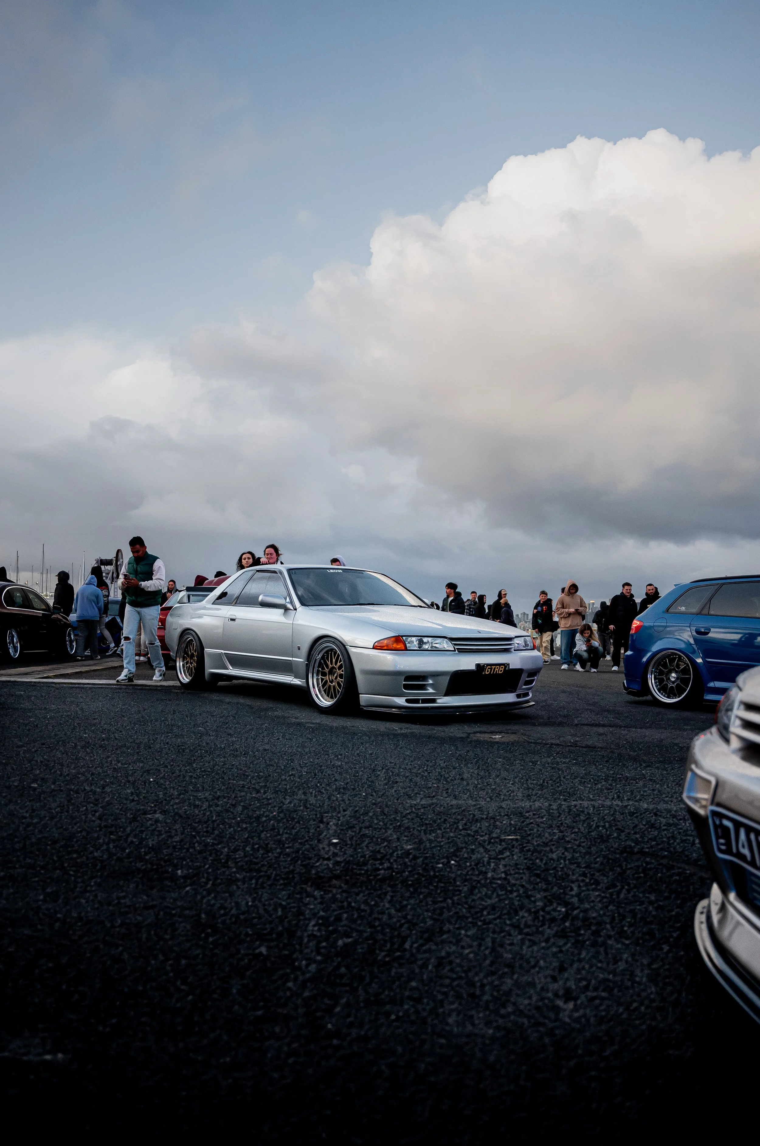 A silver sports car with gold rims parked at a car meet, with several people and additional cars in the background under a cloudy sky.