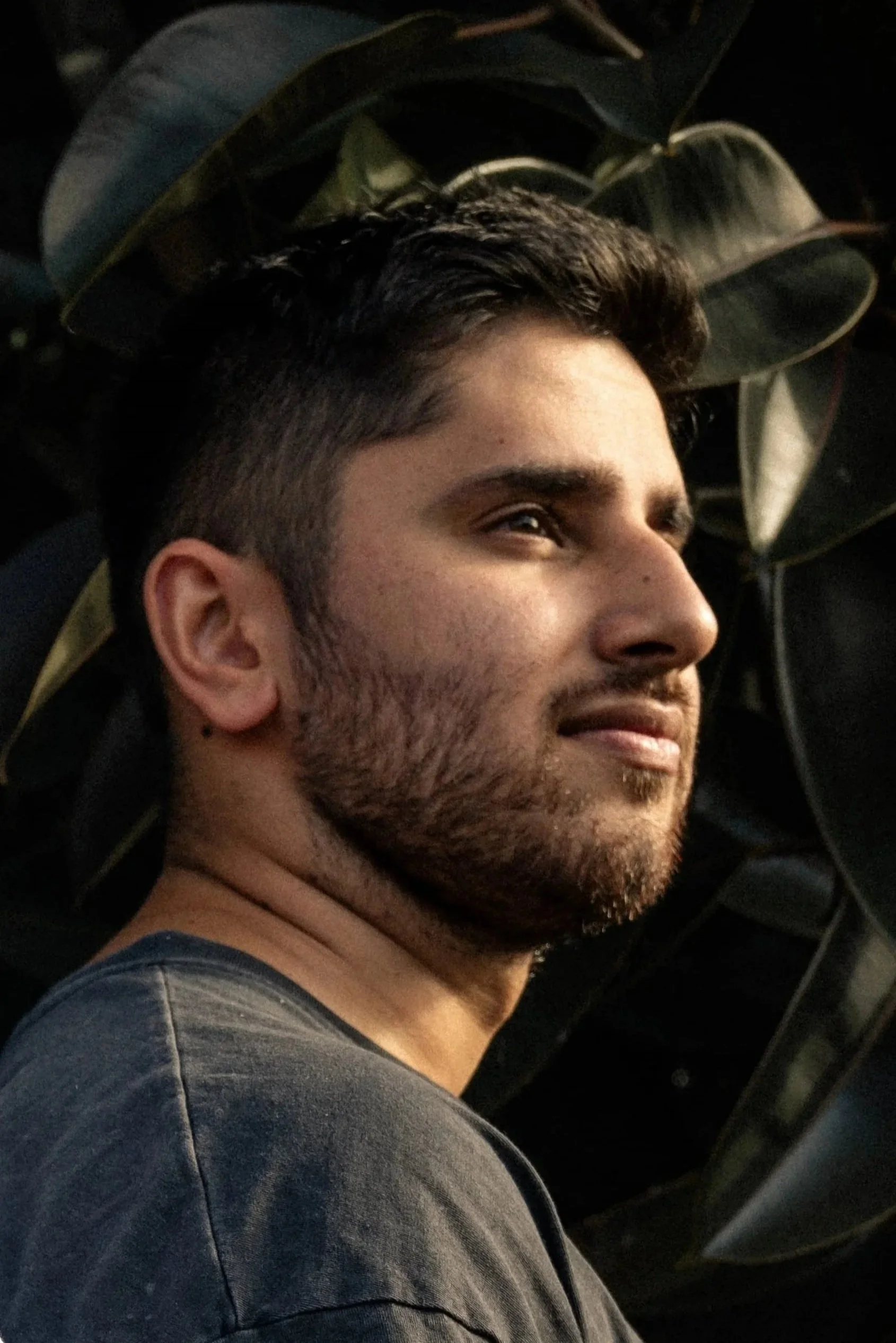 A young man with dark hair and facial hair smiling slightly, looking to the side, with large dark green leaves in the background.