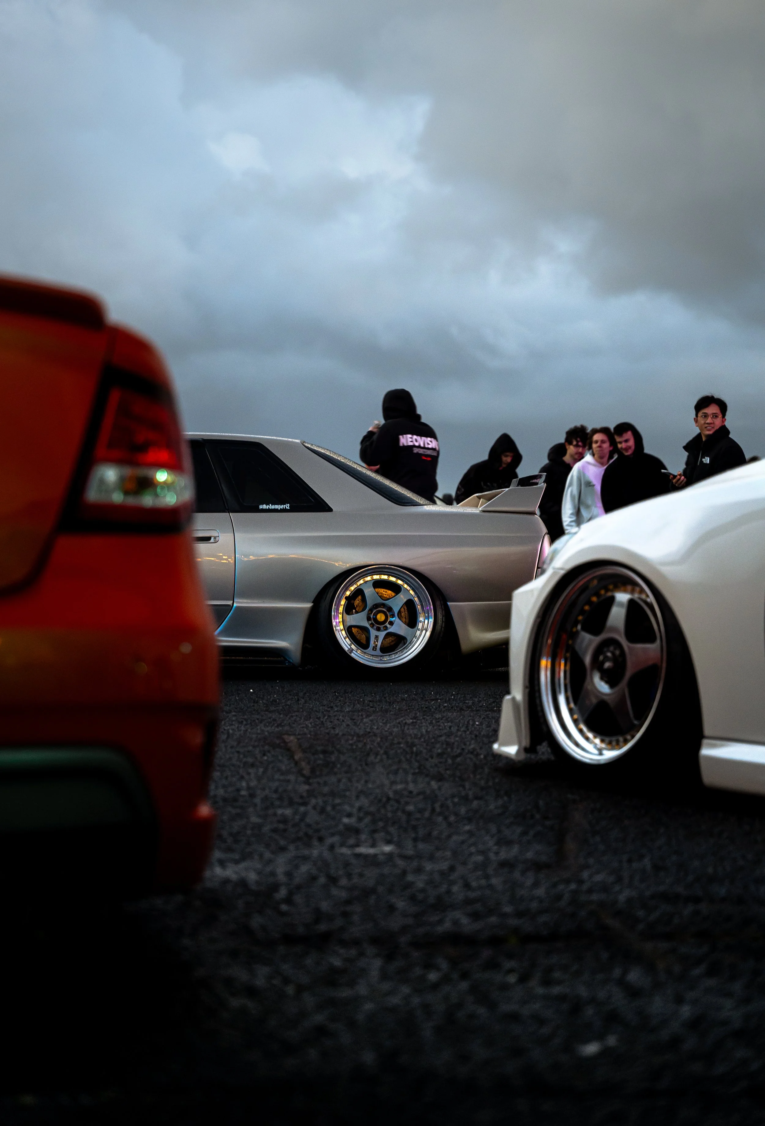 A group of people standing near modified sports cars on a dark, cloudy day, with a red vehicle partially visible in the foreground.