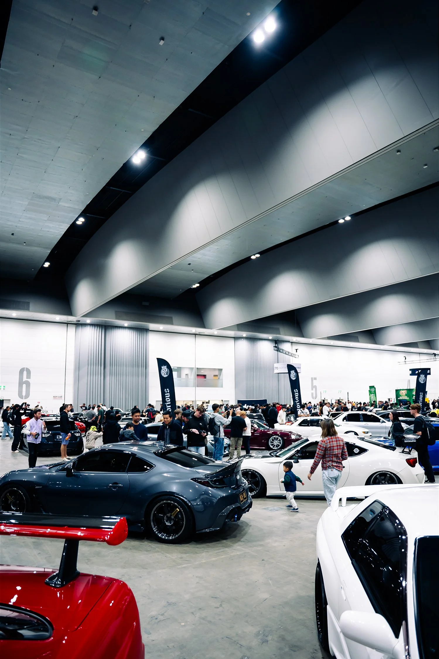 An indoor car show with various luxury and sports cars on display, crowded with people walking around and observing the vehicles.