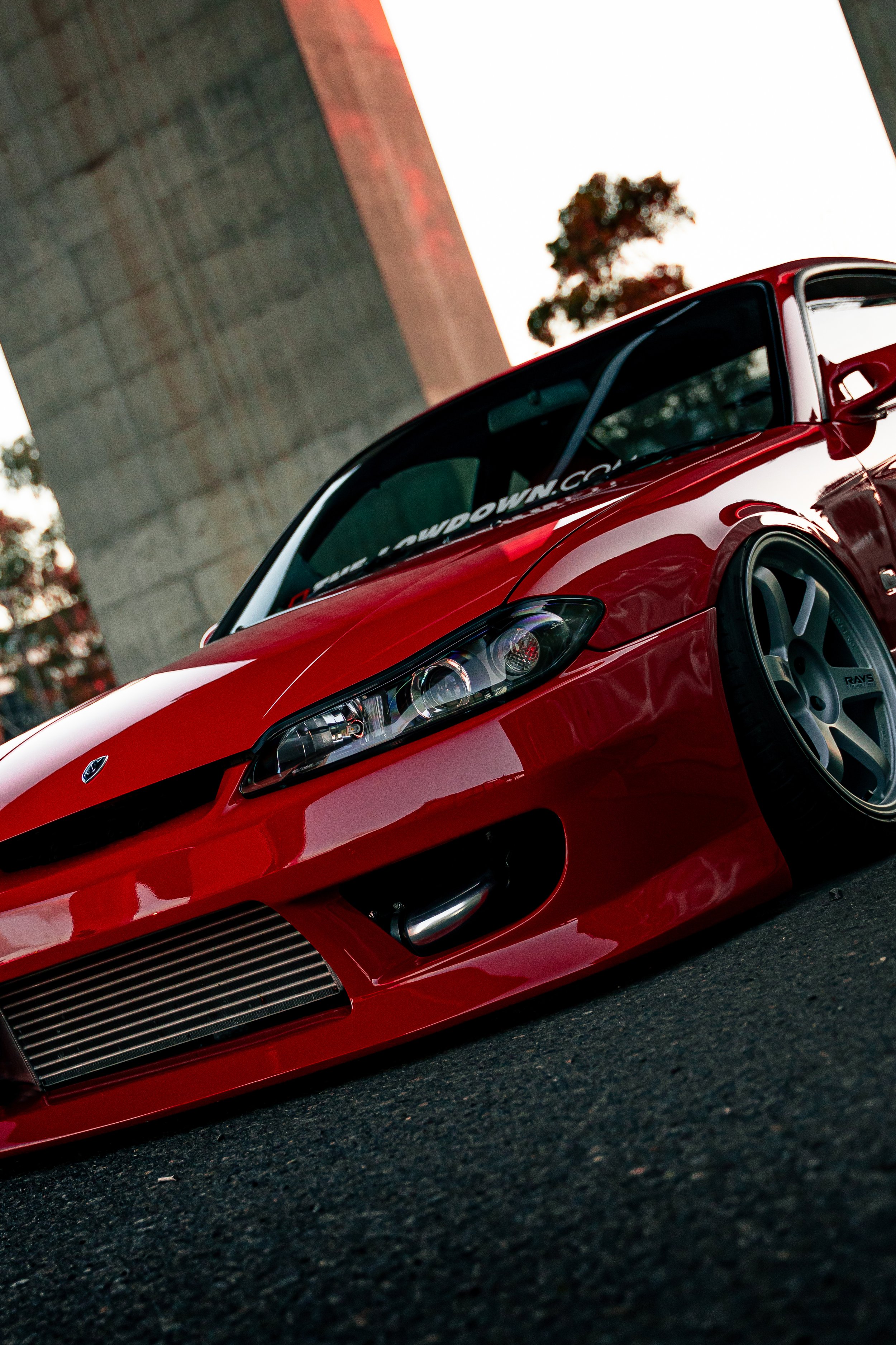 A close-up of a red sports car parked on a street, with a large concrete bridge and trees in the background during sunset.