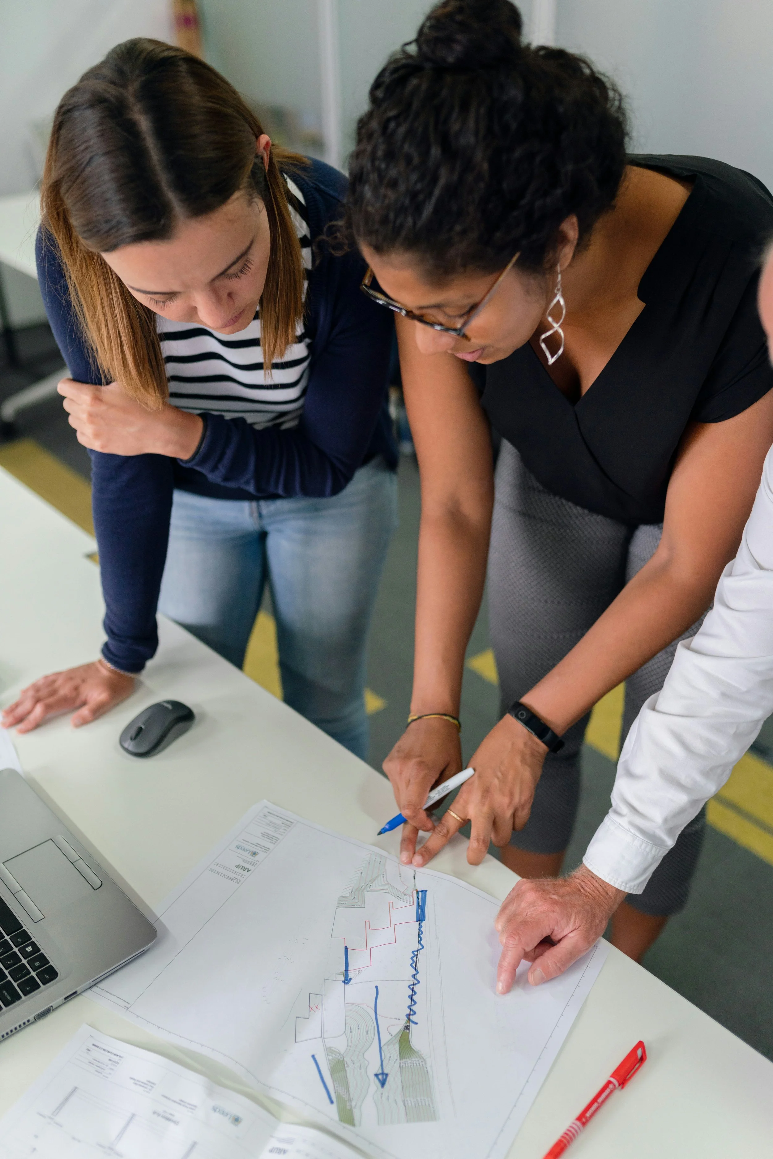 Two women and a man are examining a large map or blueprint on a table, pointing at it, with a laptop, a mouse, and a red pen nearby.