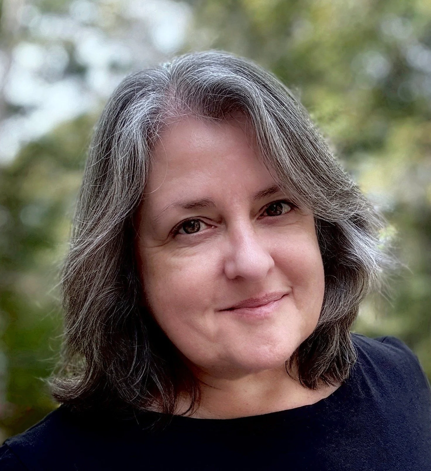 A close-up portrait of a woman with shoulder-length gray hair, smiling, outdoors with blurred green foliage in the background.