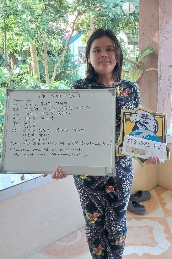 A woman in a floral outfit holding a whiteboard with handwritten text and a cartoon lion sign with Korean writing, standing near a window with trees outside.