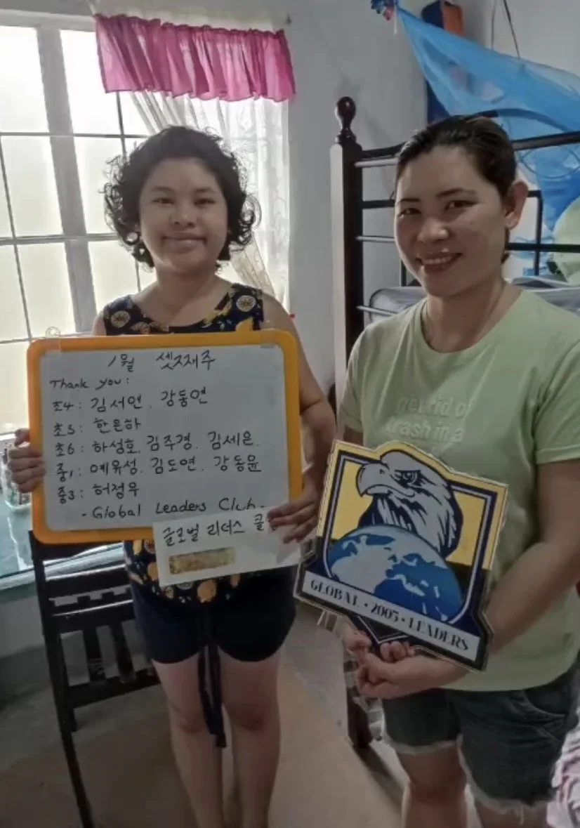 Two women standing indoors, one holding a whiteboard with Korean writing and a thank you message, and the other holding a sign with an eagle and the words 'Global Leaders Club'.