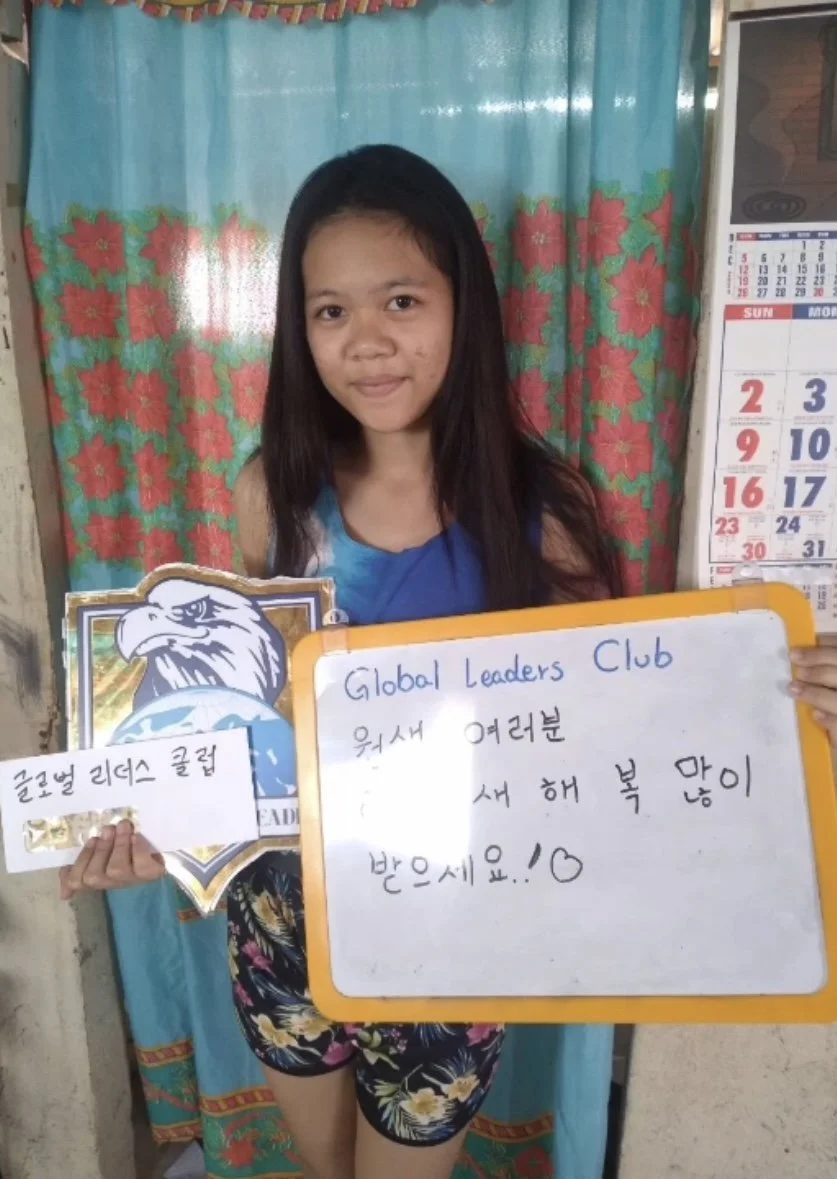 A young girl inside her home holding a whiteboard with handwritten message and a sign with an eagle logo. The whiteboard reads "Global Leaders Club" and includes a message in Korean. The background features a colorful floral curtain and a calendar on