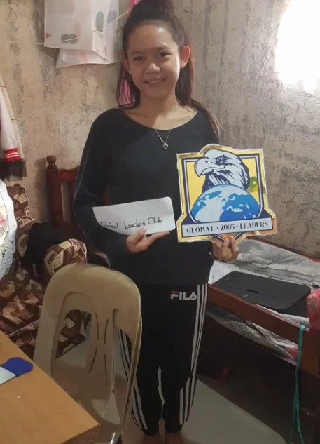 Young girl holding a sign and a logo in a room with a concrete wall, a desk with a laptop, and a plastic chair.