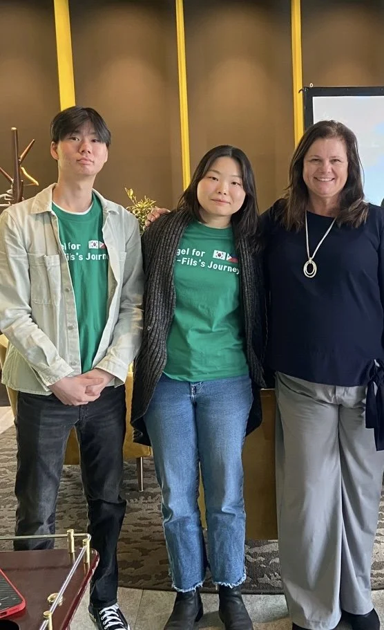 Three people standing together indoors, two young individuals wearing green shirts with white text, and an older woman in a dark top and gray pants, posing for a photo.