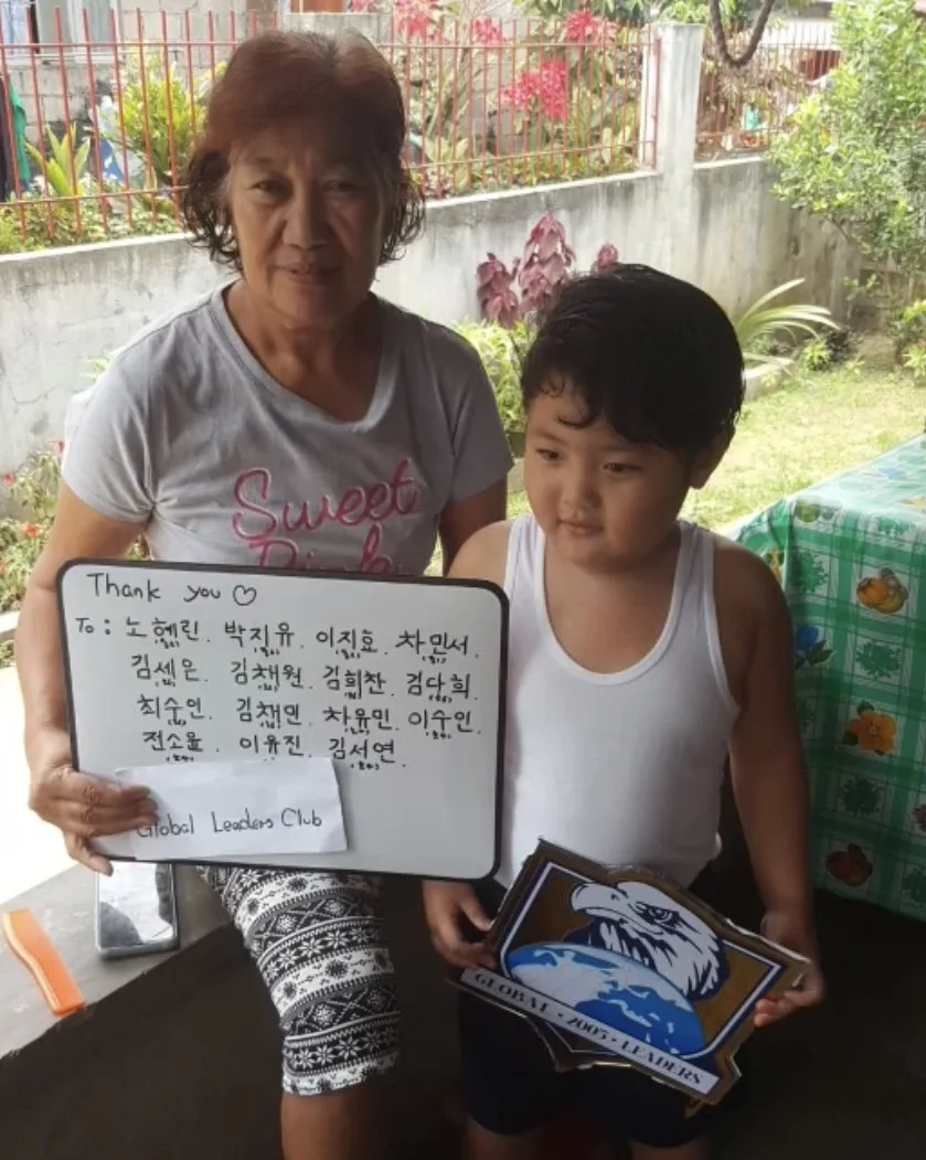 An elderly woman and a young boy standing outdoors, holding a whiteboard with handwritten message and a set of stickers or cards. The woman is wearing a gray T-shirt with pink writing, and the boy is in a white tank top. They are in a garden with pla