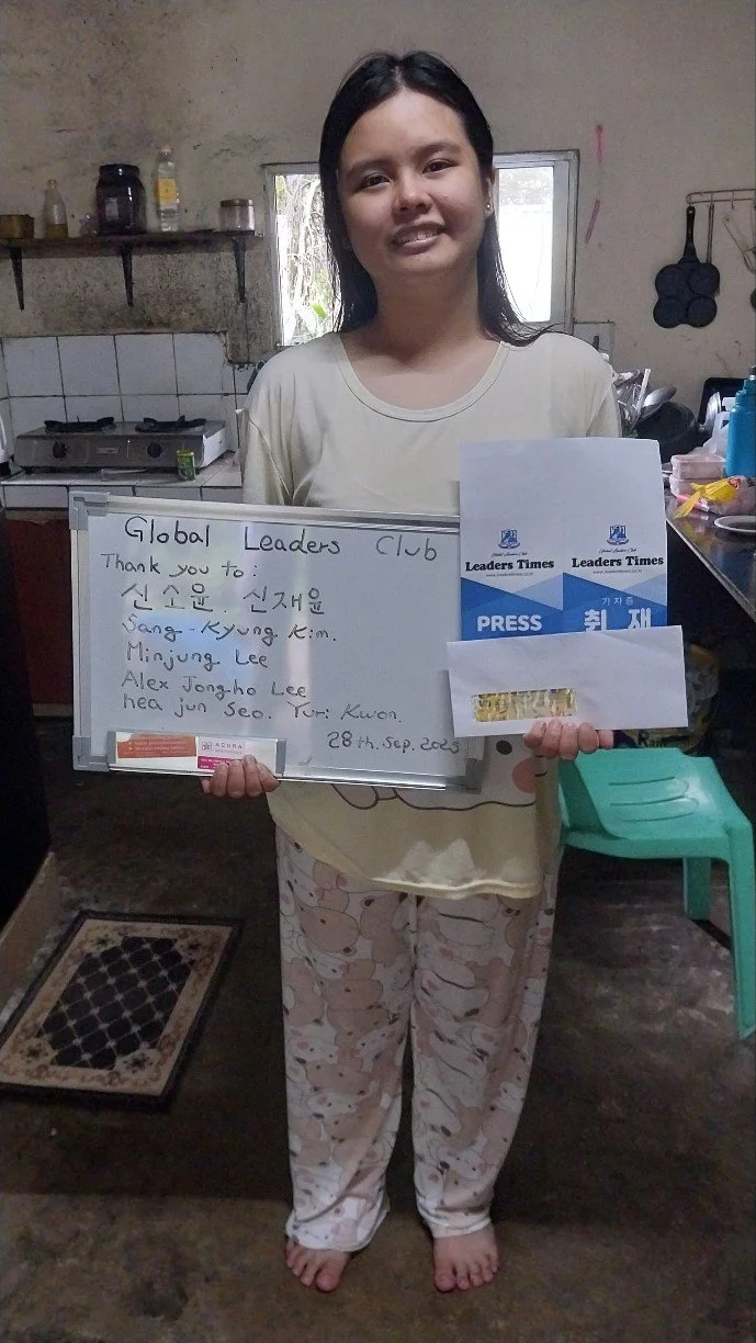A young girl standing barefoot in a kitchen, holding a whiteboard with a list of names and thank you notes, and a newspaper clipping titled 'Press.'