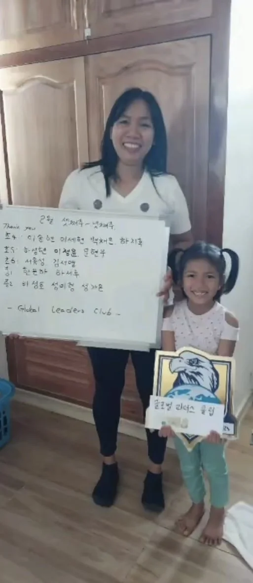 A woman and a young girl standing inside a room with wooden cabinets. The woman is holding a large whiteboard with writing in Korean, and the girl is holding a sign with an eagle and blue background. Both are smiling at the camera.