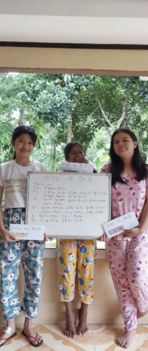 Three young girls standing together indoors, two holding white envelopes, with a whiteboard displaying handwritten notes in the middle, smiling and looking at the camera, with a window showing green trees in the background.