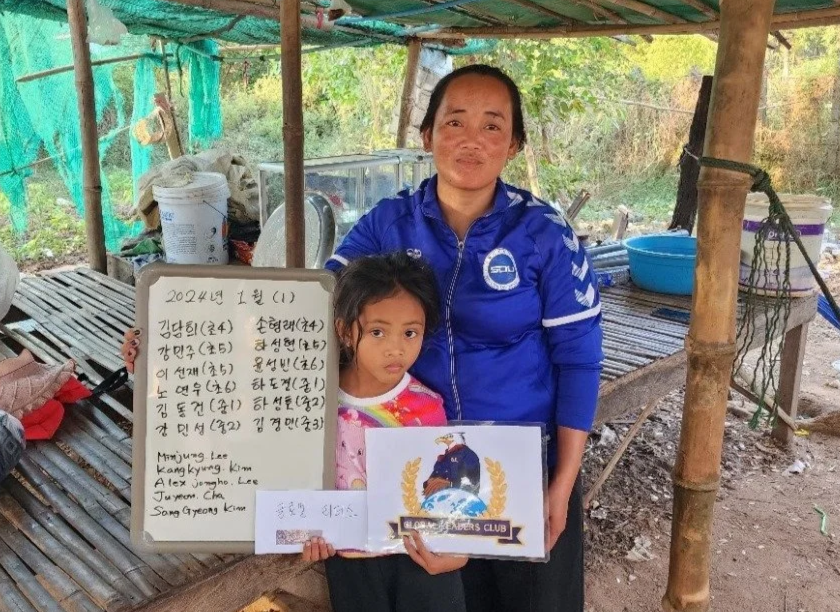 A woman in a blue jacket and a young girl holding a certificate and a whiteboard with handwritten text, standing inside a rustic outdoor shelter with bamboo and green tarps.