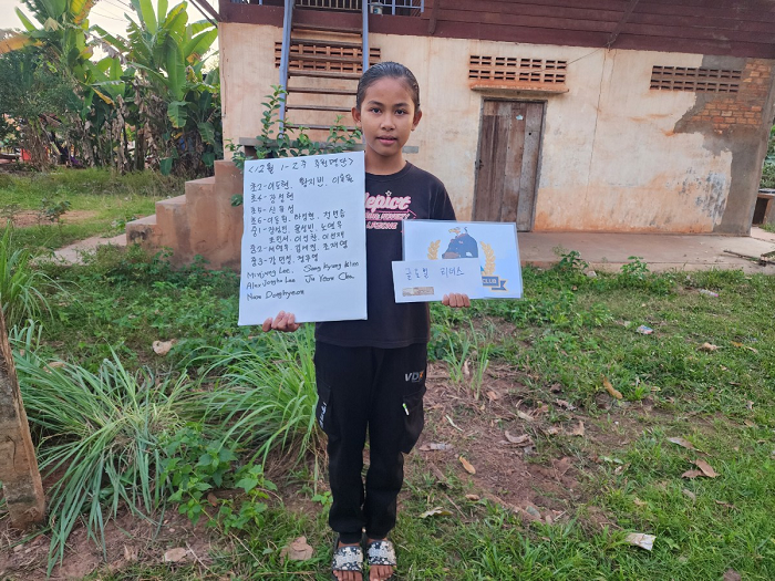 A young girl standing outdoors on grass holding a whiteboard with writing and a poster showing a blue cartoon character.